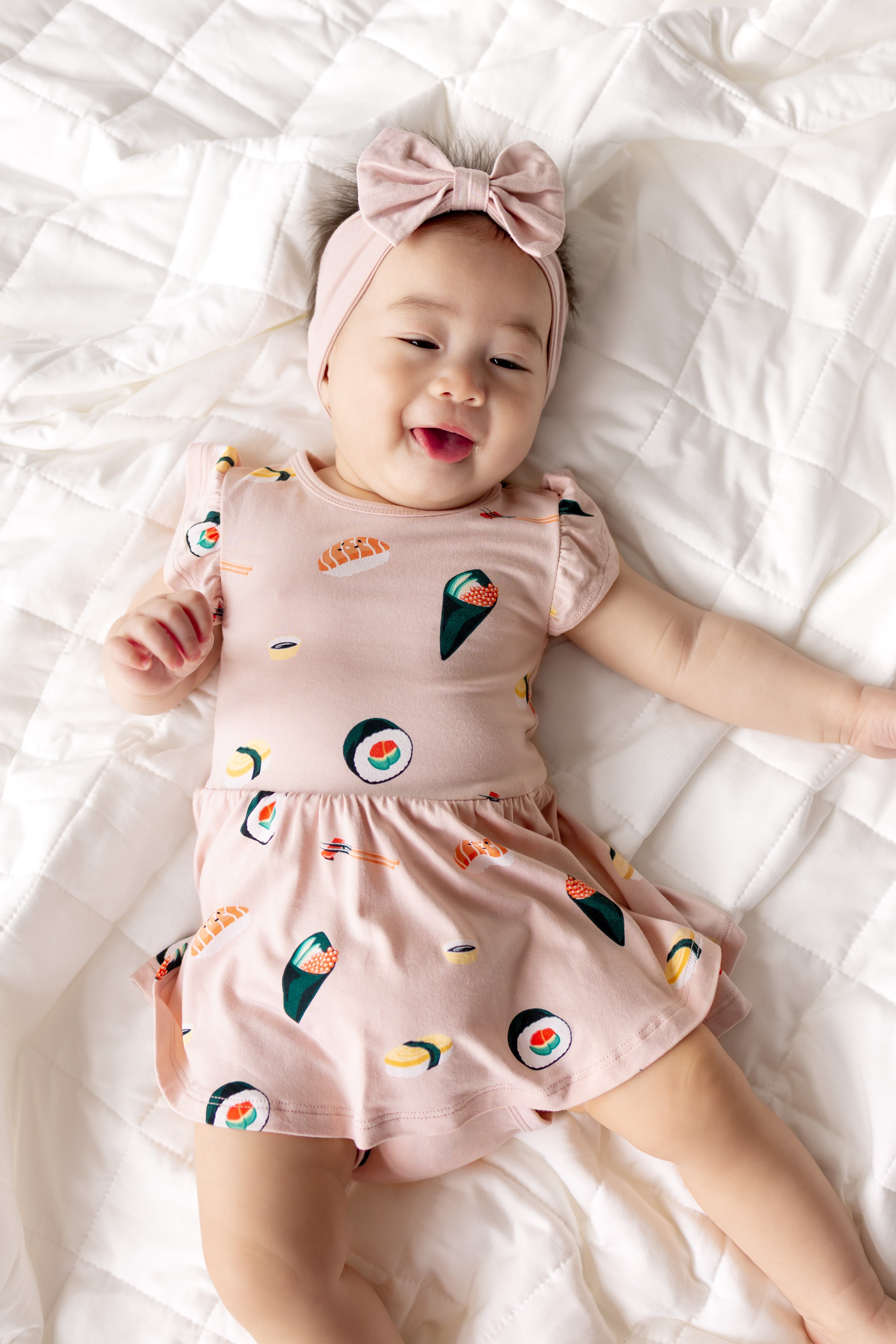 Baby lying on white sheet wearing Pink Twirl Bodysuit Dress with Sushi print and pink headband