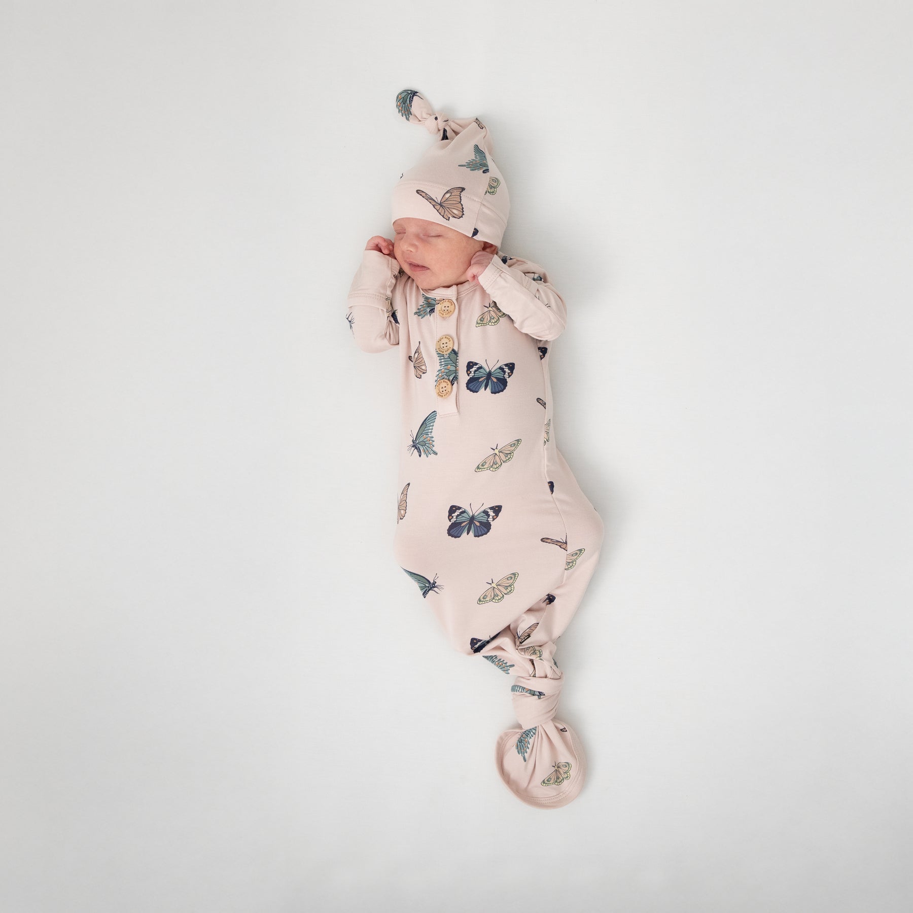 Sleeping newborn laying on a white blanket wearing the Knotted Gown with Hat Set in Blush Butterfly knotted at the bottom