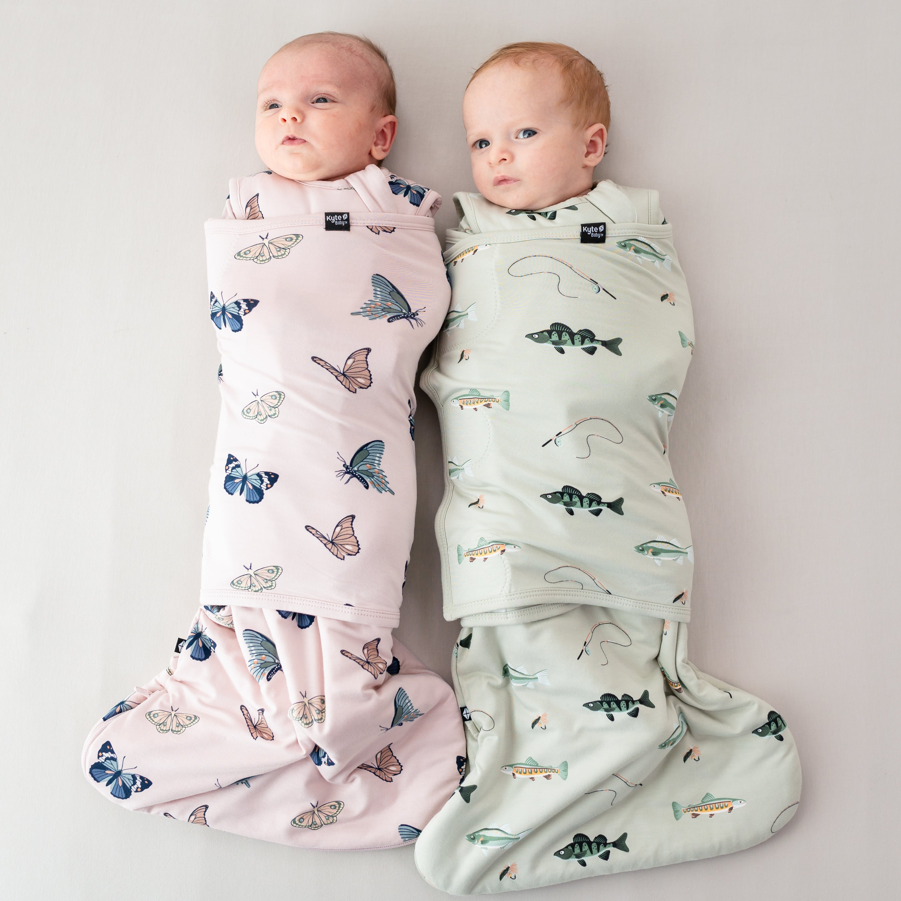 Two babies laying down swaddled in Sleep bag swaddlers in Blush Butterfly and Fishing 