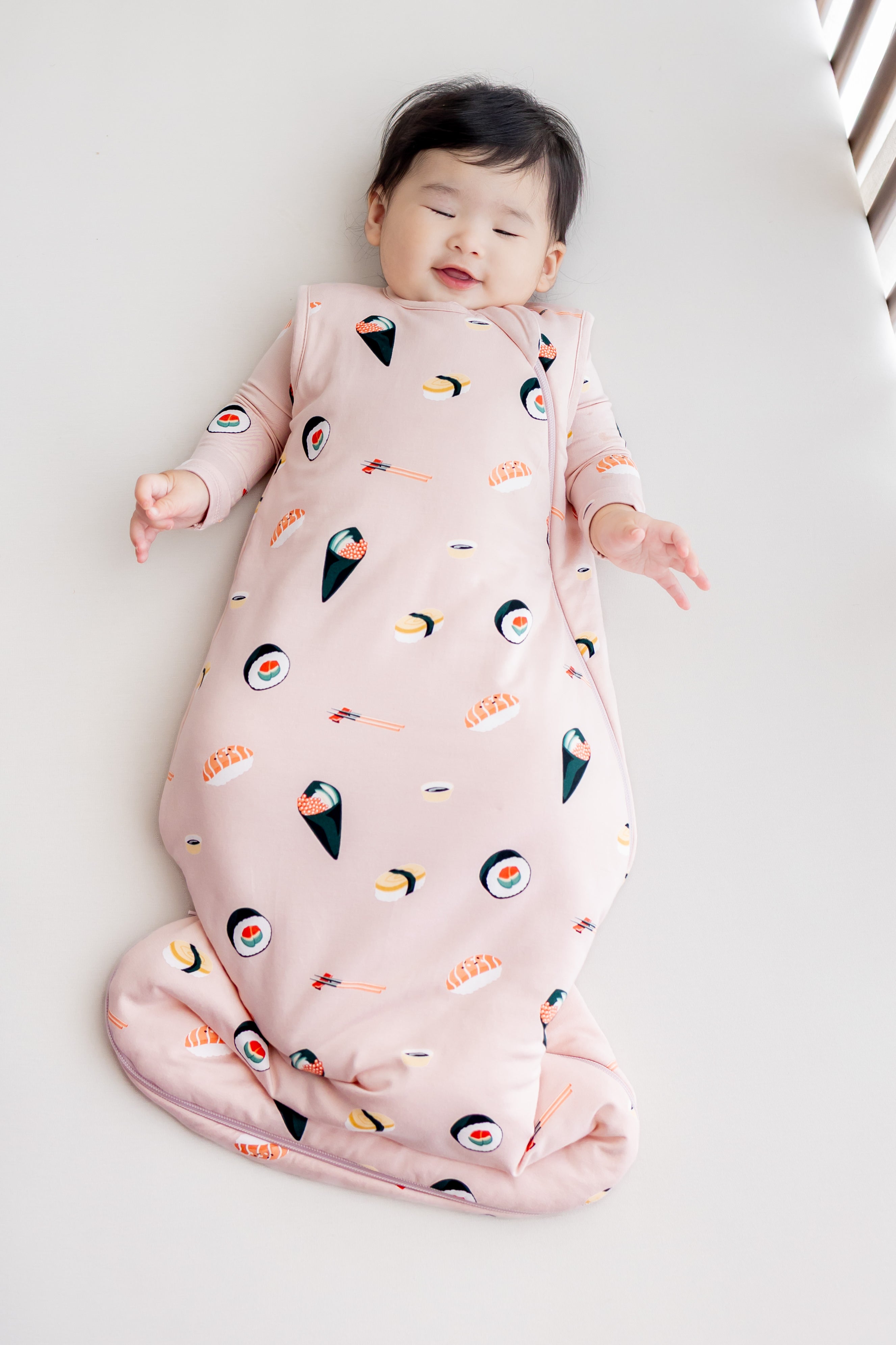 Baby laying down in a crib with white sheets in lush sushi sleep bag