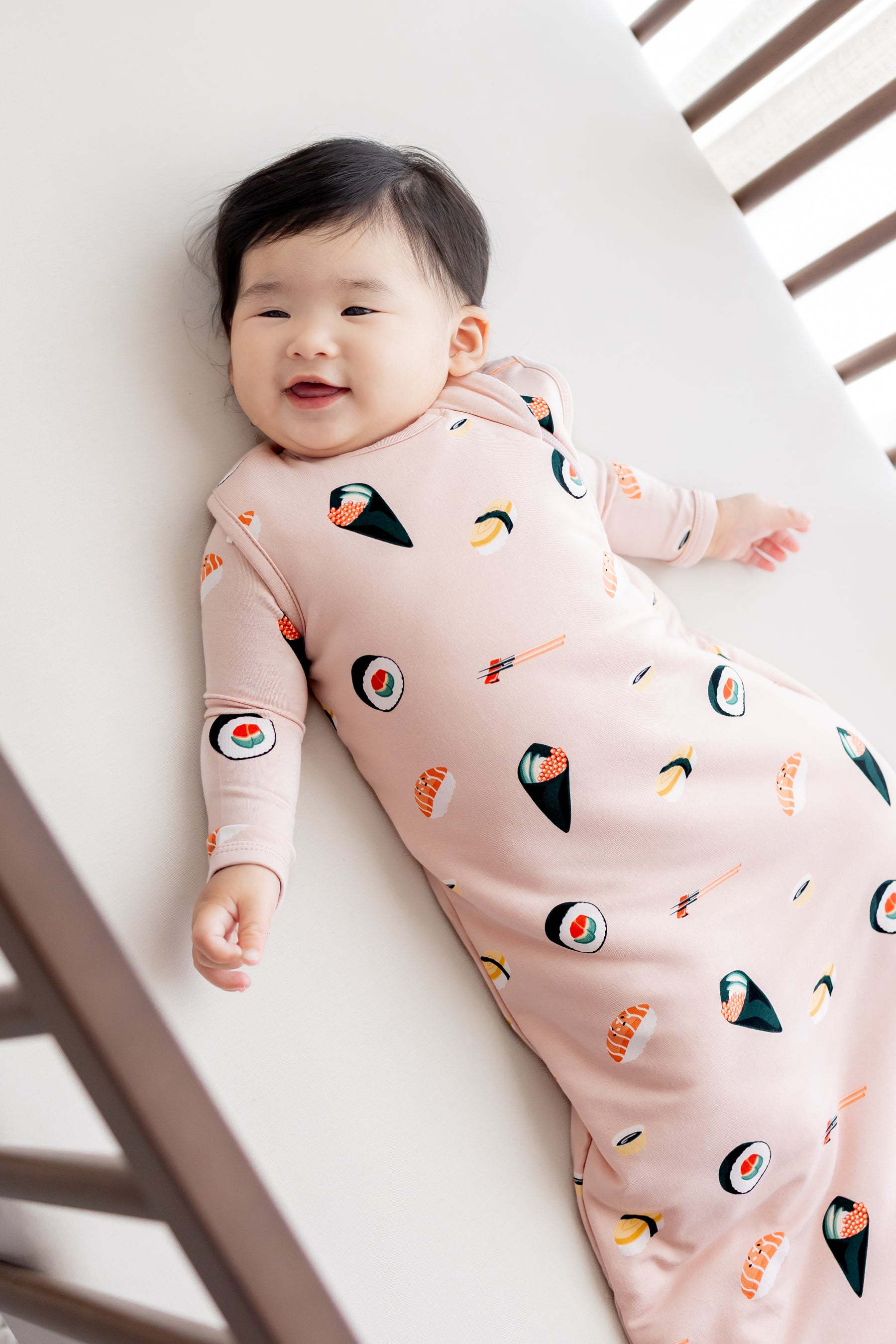 Baby laying in crib with white sheets smiling in blush sushi sleep bag