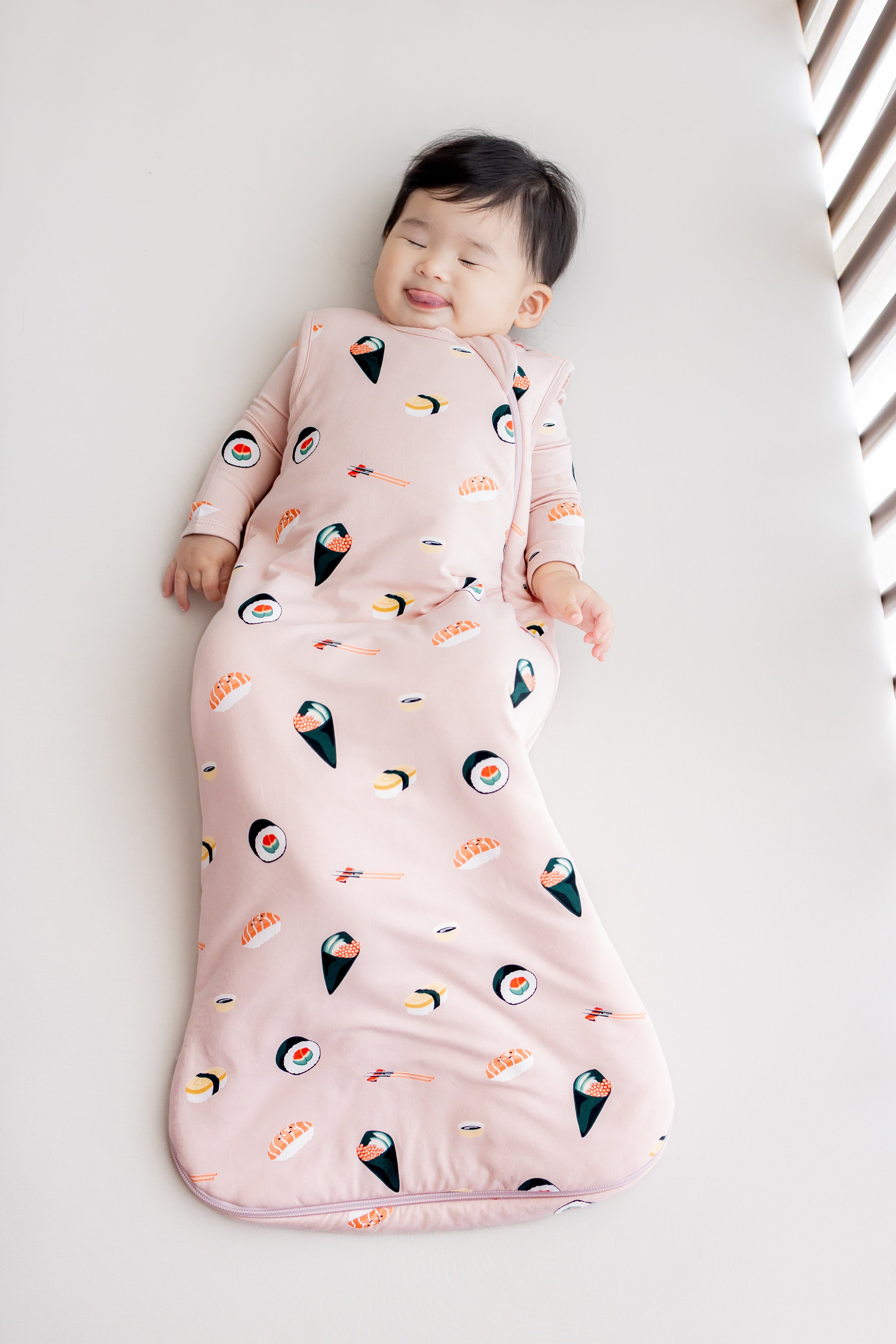 Baby laying in crib with white sheets in blush sushi sleep bag