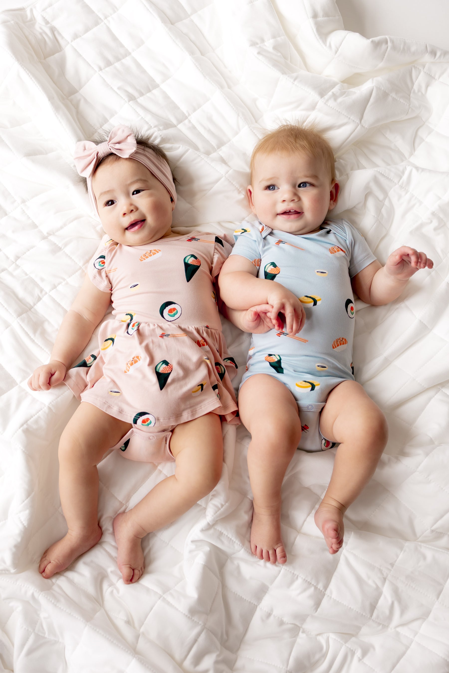 Two babies lying on white sheet wearing a pink twirl bodysuit and blue short sleeve bodysuit with sushi print on them