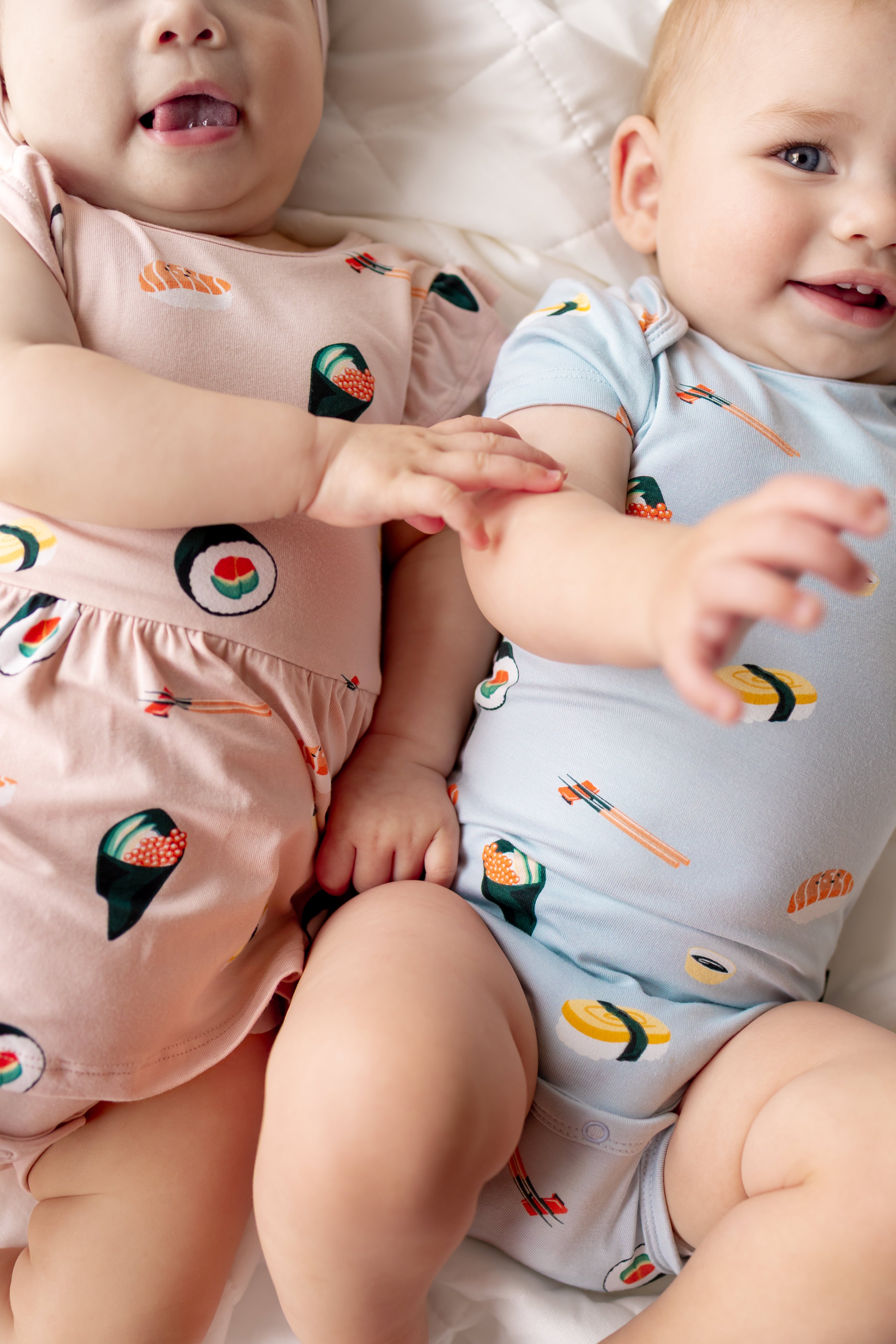 Two babies lying on white sheet wearing a pink twirl bodysuit and blue short sleeve bodysuit with sushi print on them