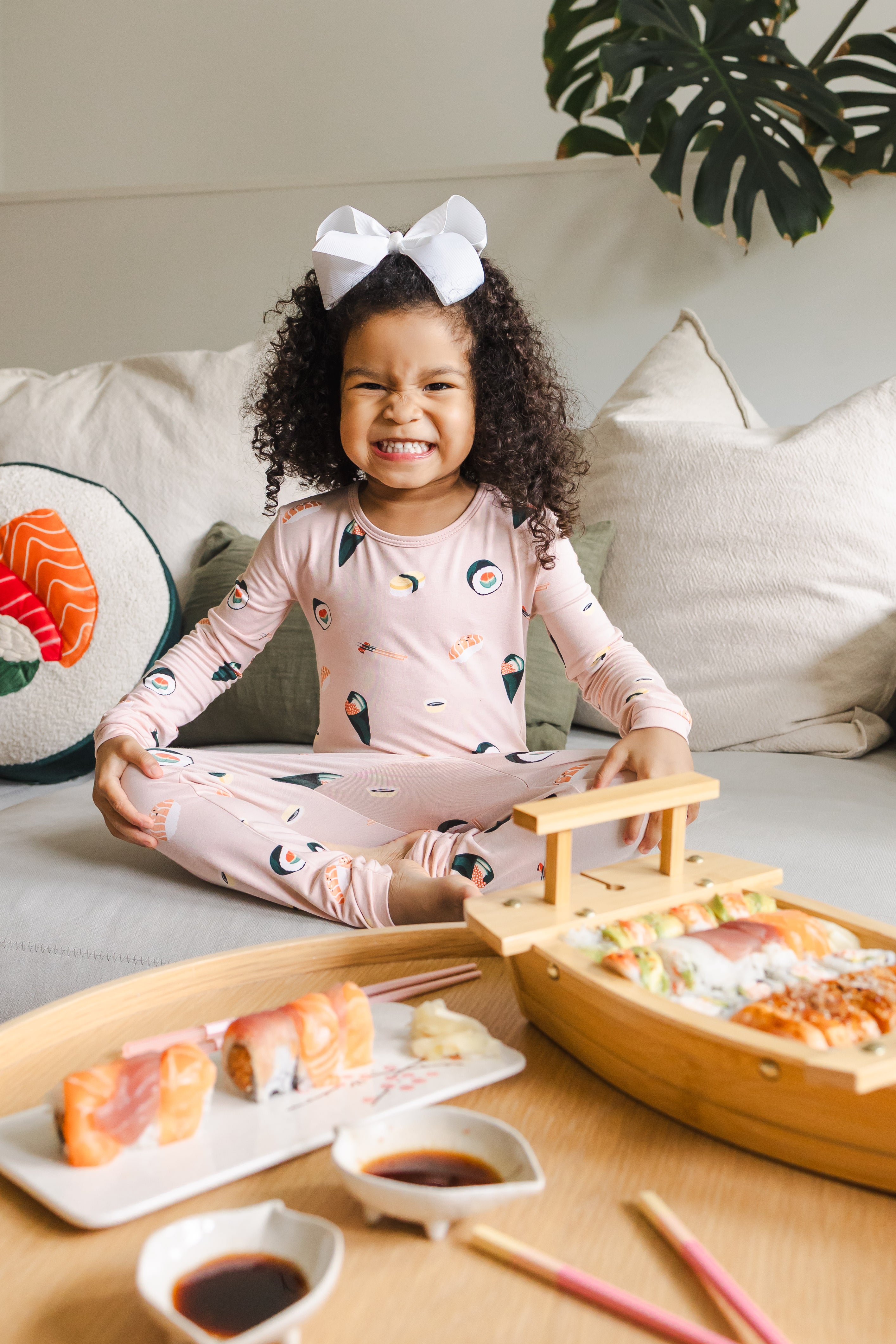 Child sitting crisscross on a white couch  wearing blush sushi pajama set and white bow with sushi on a table in front of her