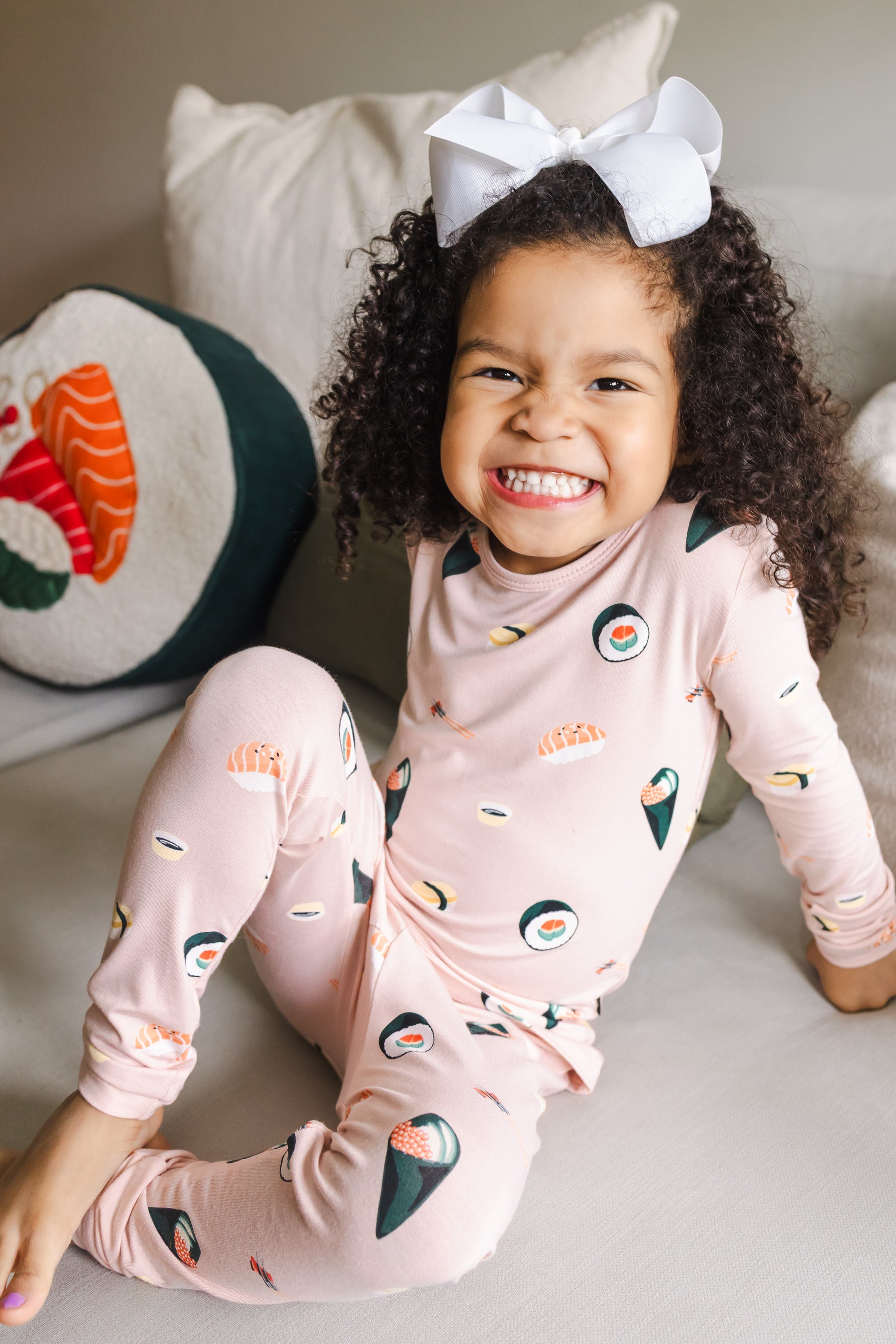Child smiling sitting on a white couch  with one leg up wearing blush sushi pajama set with a white bow