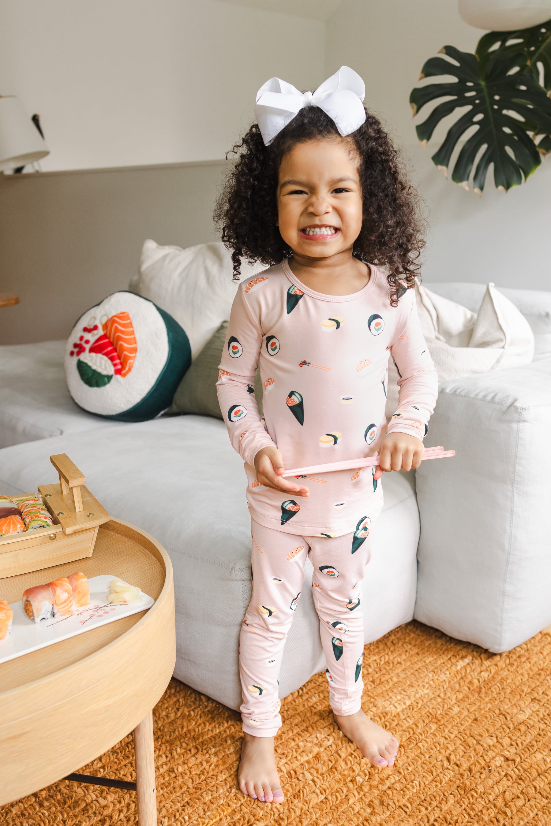 Child smiling with chop sticks in her hand wearing blush sushi pajama set with a white couch in the background