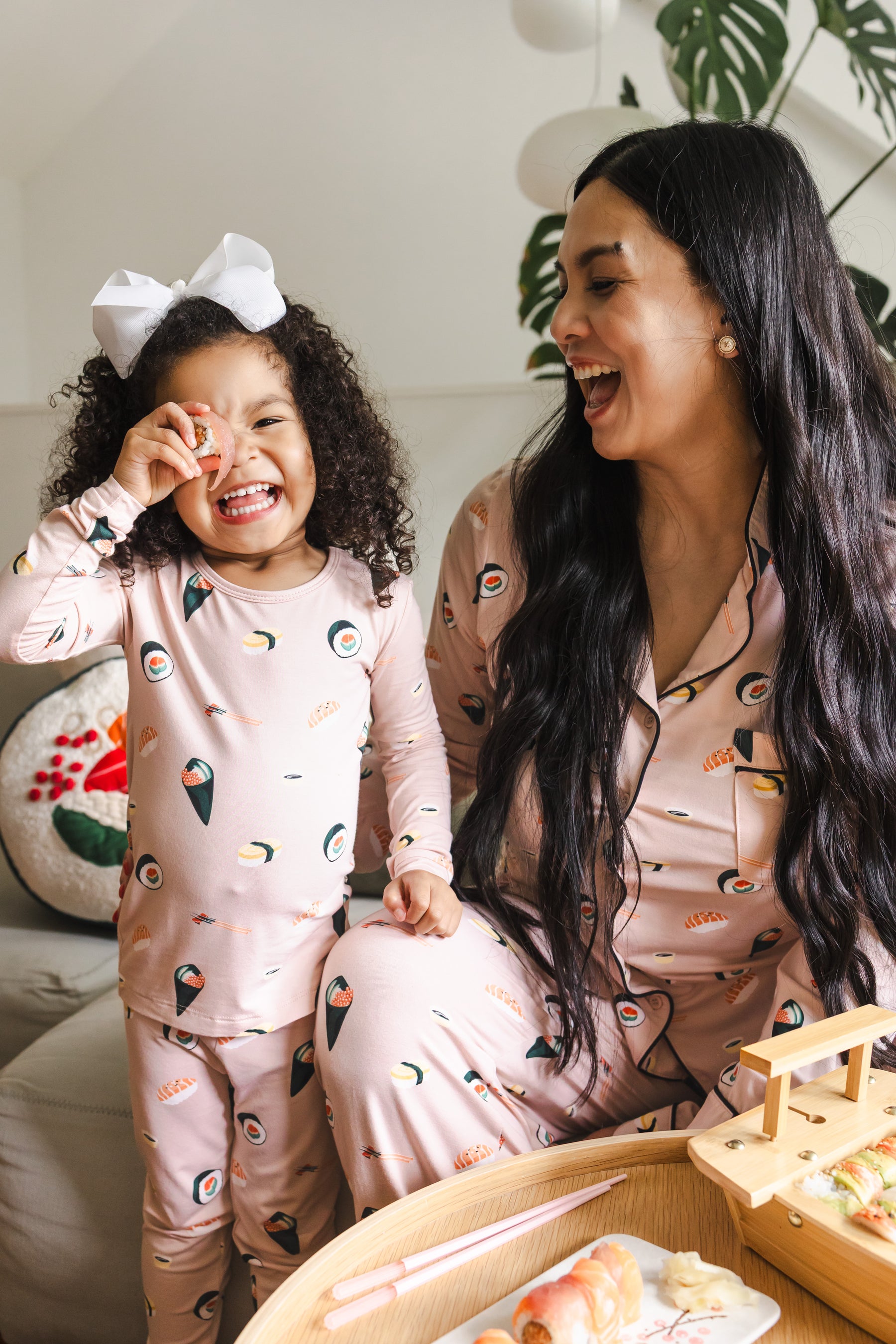 A woman and a young child wearing matching light pink pajamas with a playful sushi print, sitting together in a cozy indoor setting.