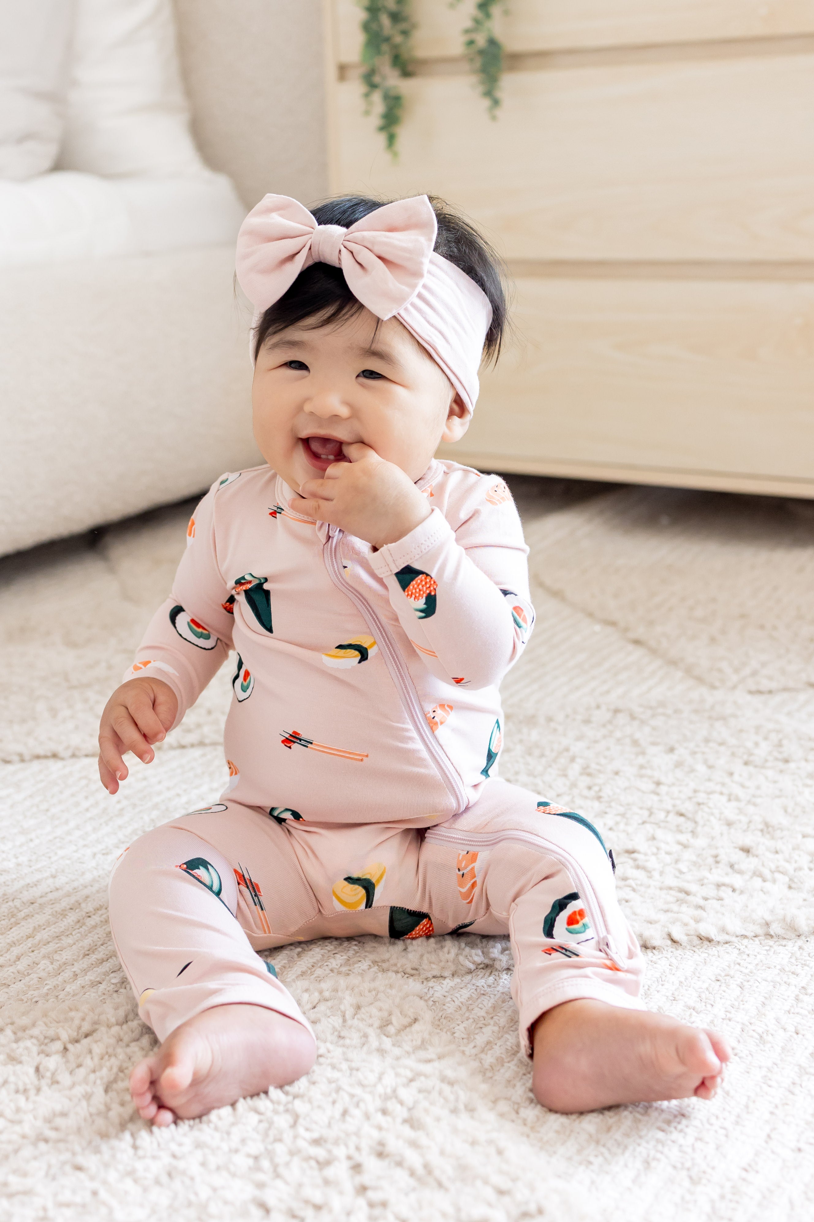 Baby sitting up with her hand in her mouth in blush sushi zipper romper and pink bow
