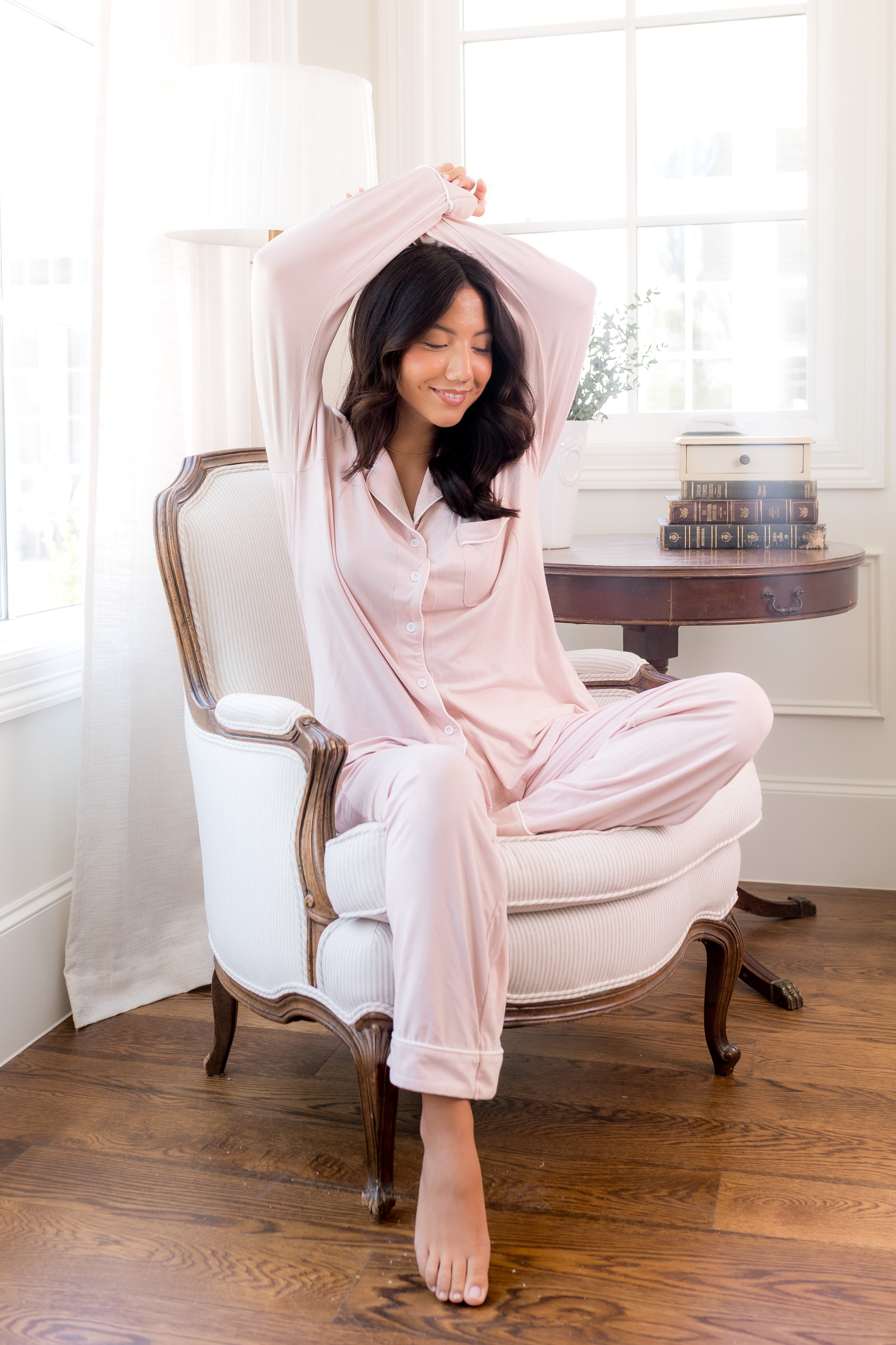 Female sitting on a chair with arms above her head wearing pink pajamas with white trim