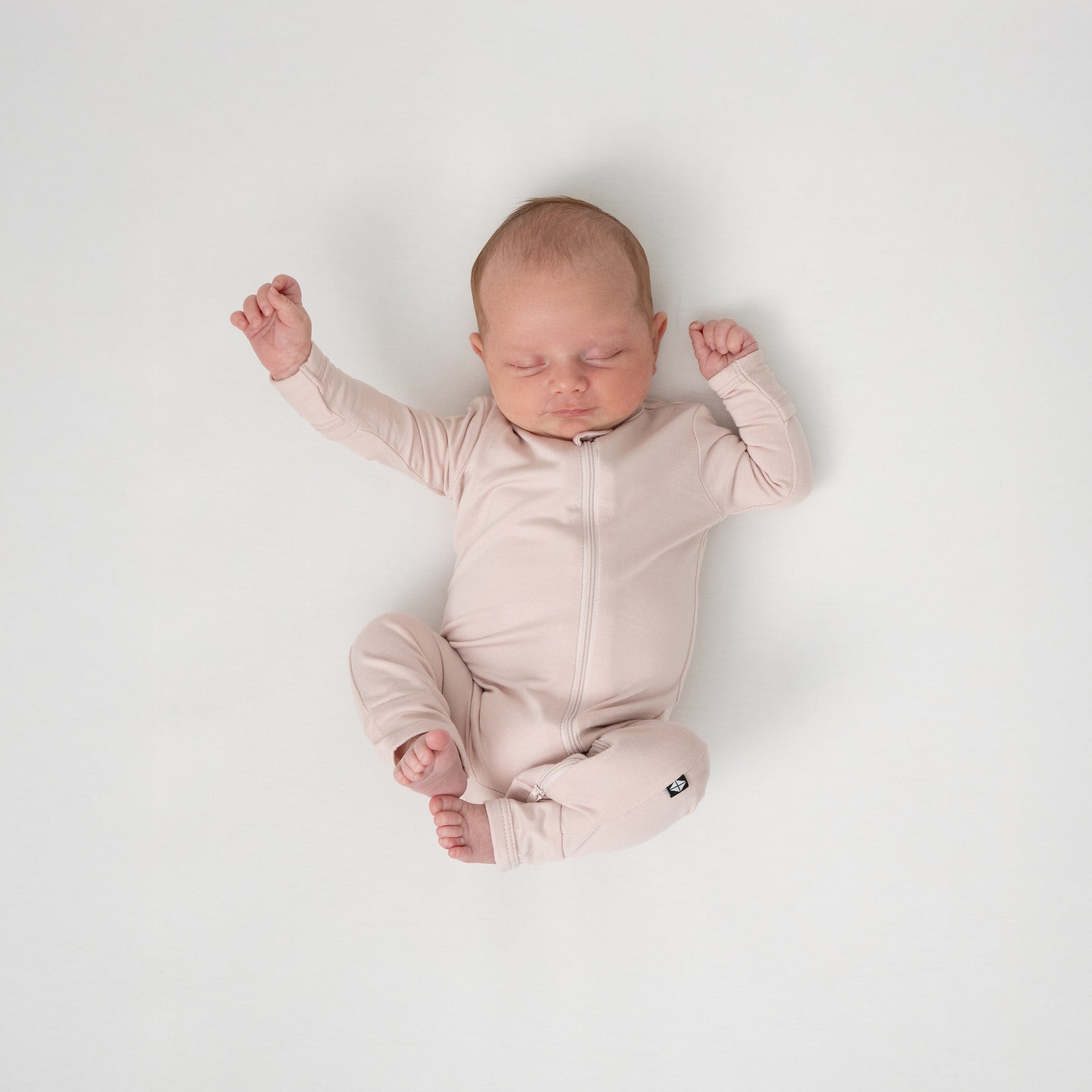 Sleeping newborn laying down on a white blanket with her legs up wearing the Zippered Romper in Blush