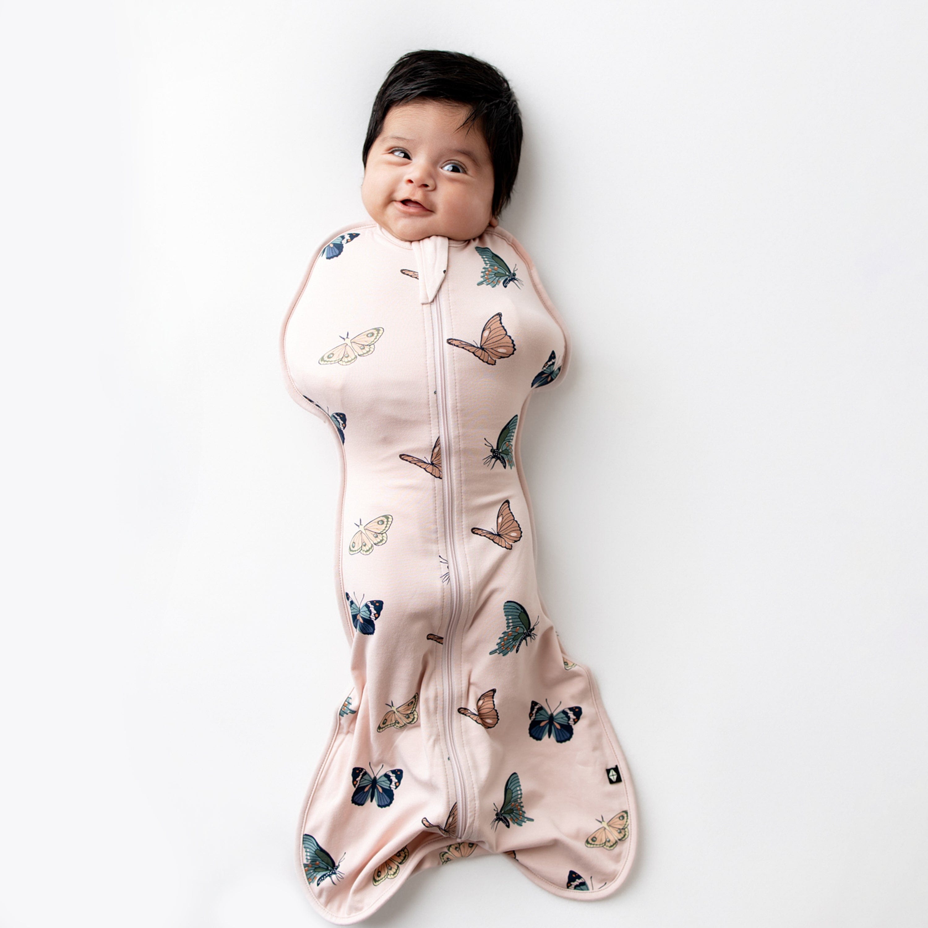 Infant laying on a white hard surface wearing the Swaddle Bag in Blush Butterfly 0.5