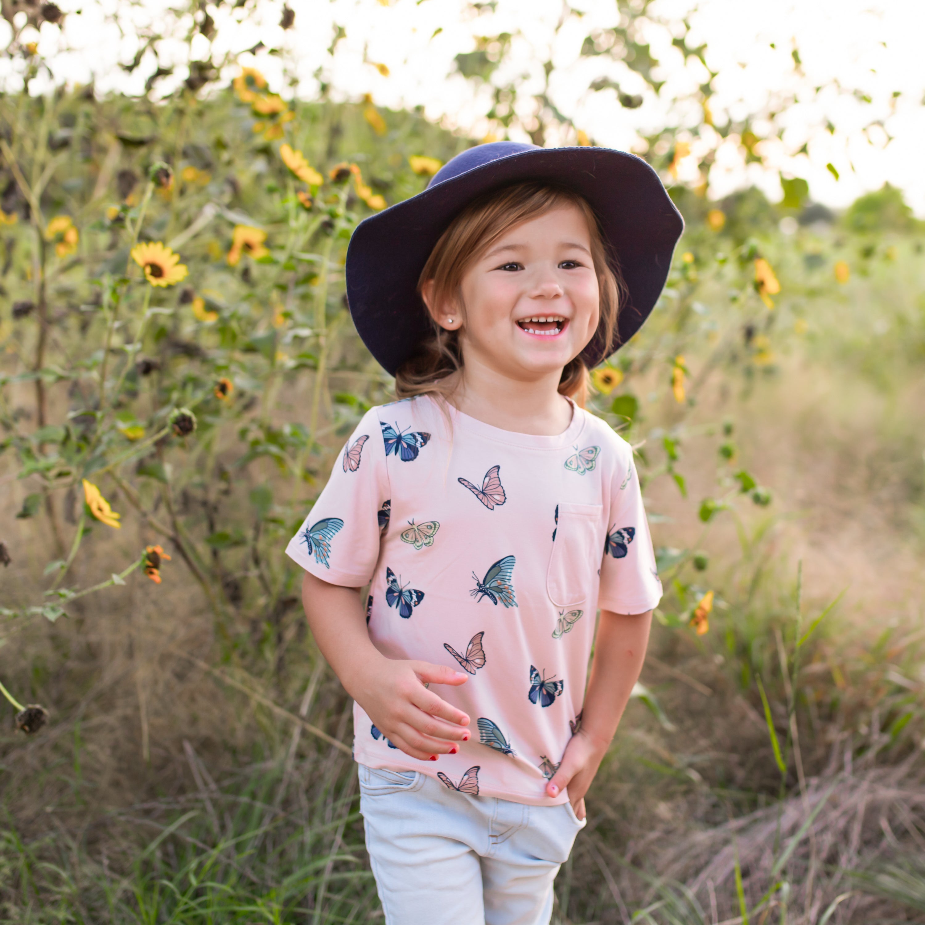 Child wearing a butterfly-patterned shirt and wide-brimmed hat standing in a sunflower field.