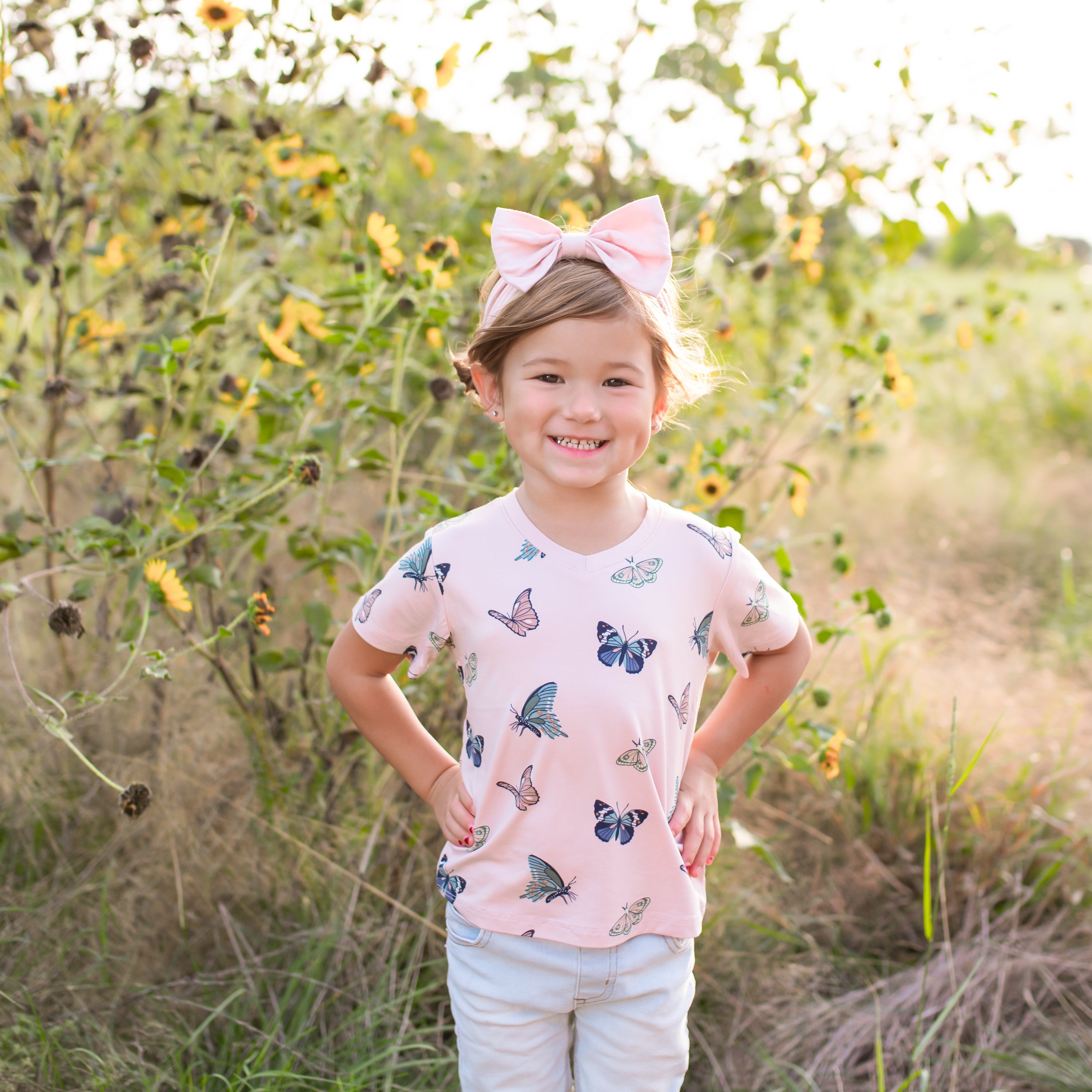 Young girl with a pink bow standing in a sunflower field wearing a butterfly-patterned shirt.