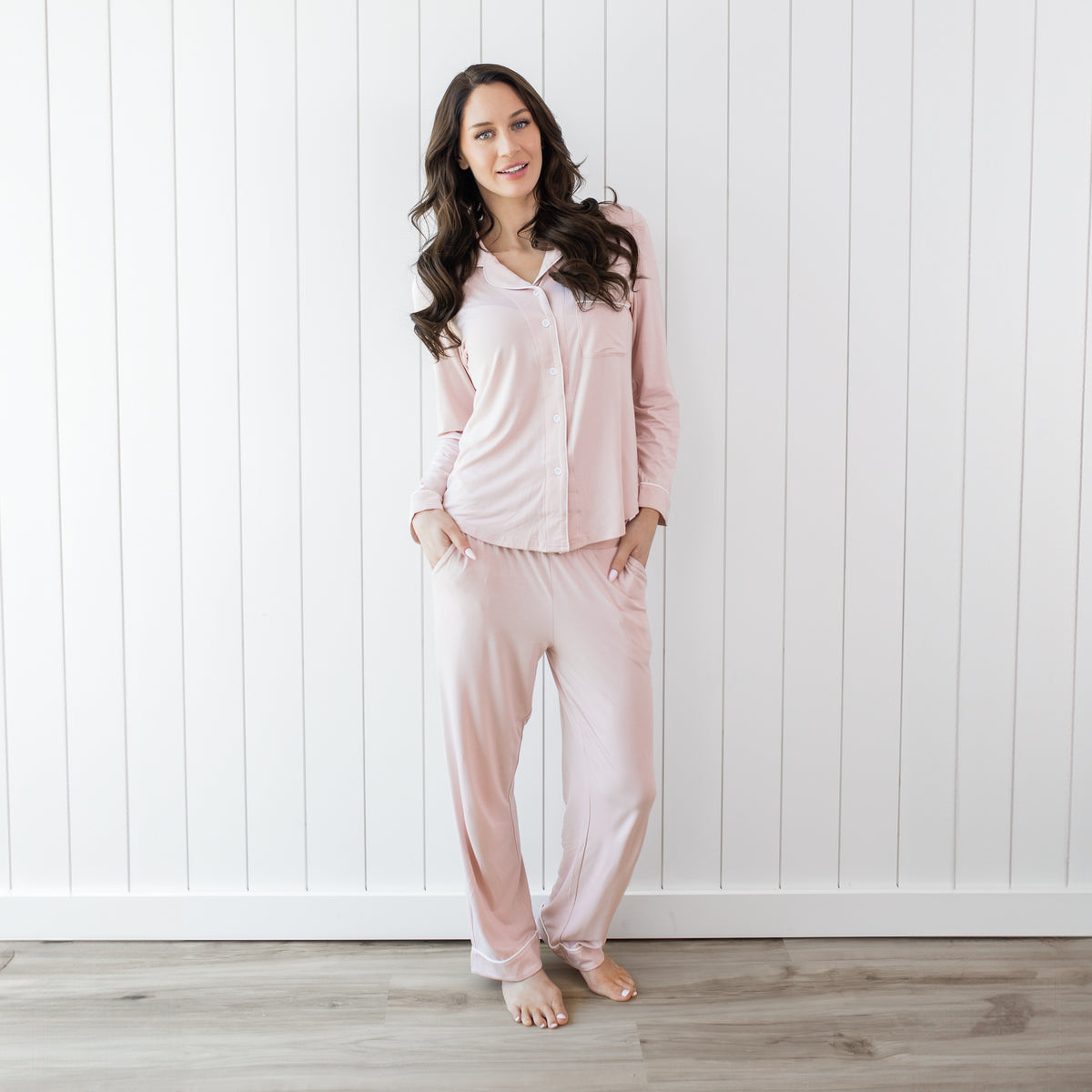 Woman wearing long sleeve button up blush pajamas with white piping against a white wooden wall backdrop