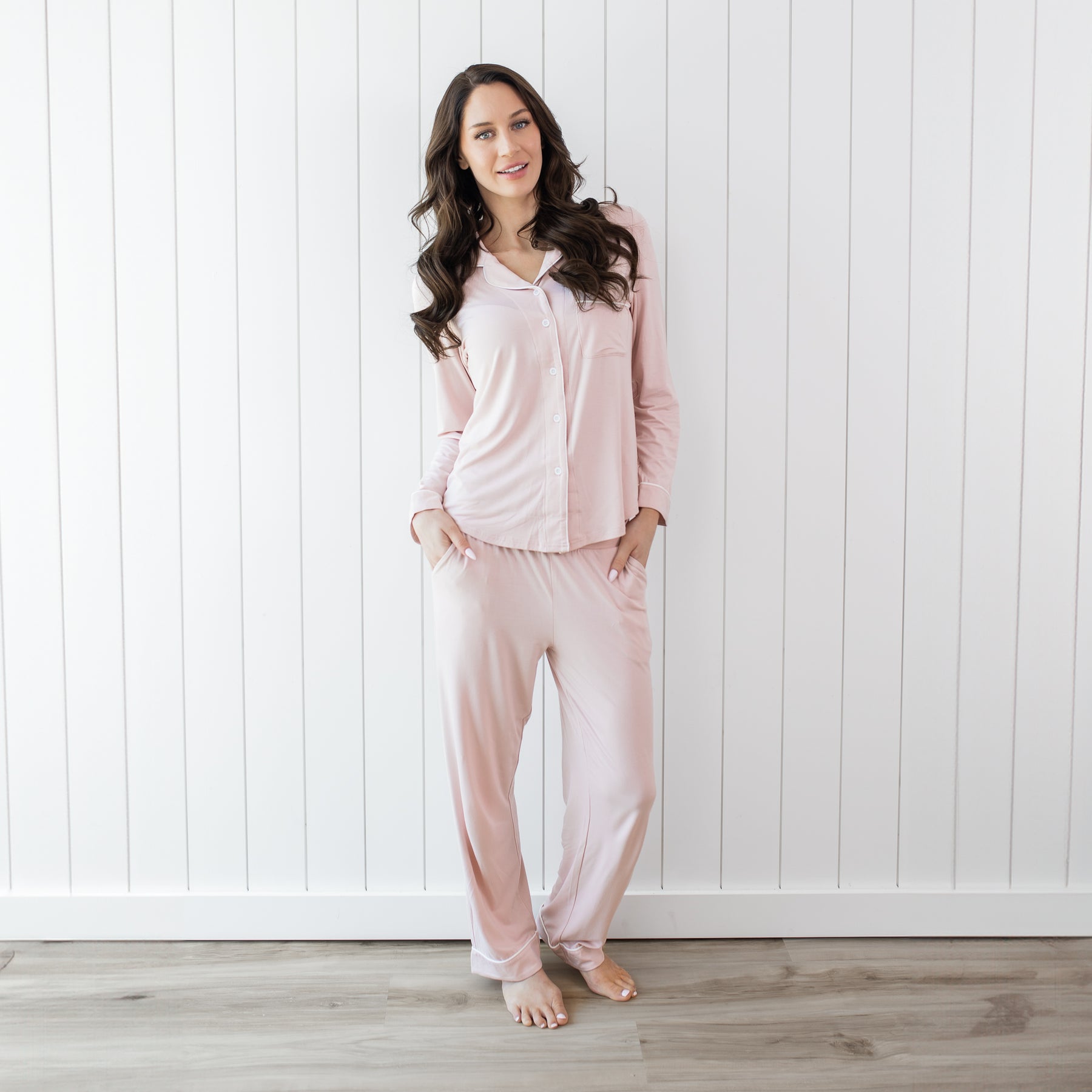Woman wearing long sleeve button up blush pajamas with white piping against a white wooden wall backdrop