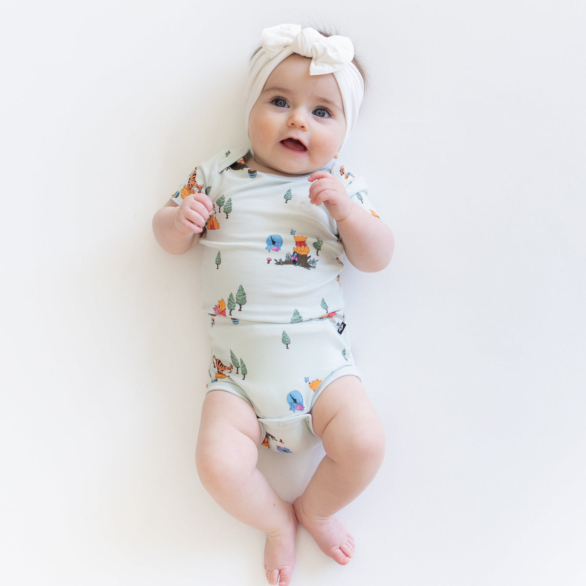 Infant girl laying on a light colored surface wearing the Bodysuit in Winnie the Pooh Hundred Acre Wood with a white bow