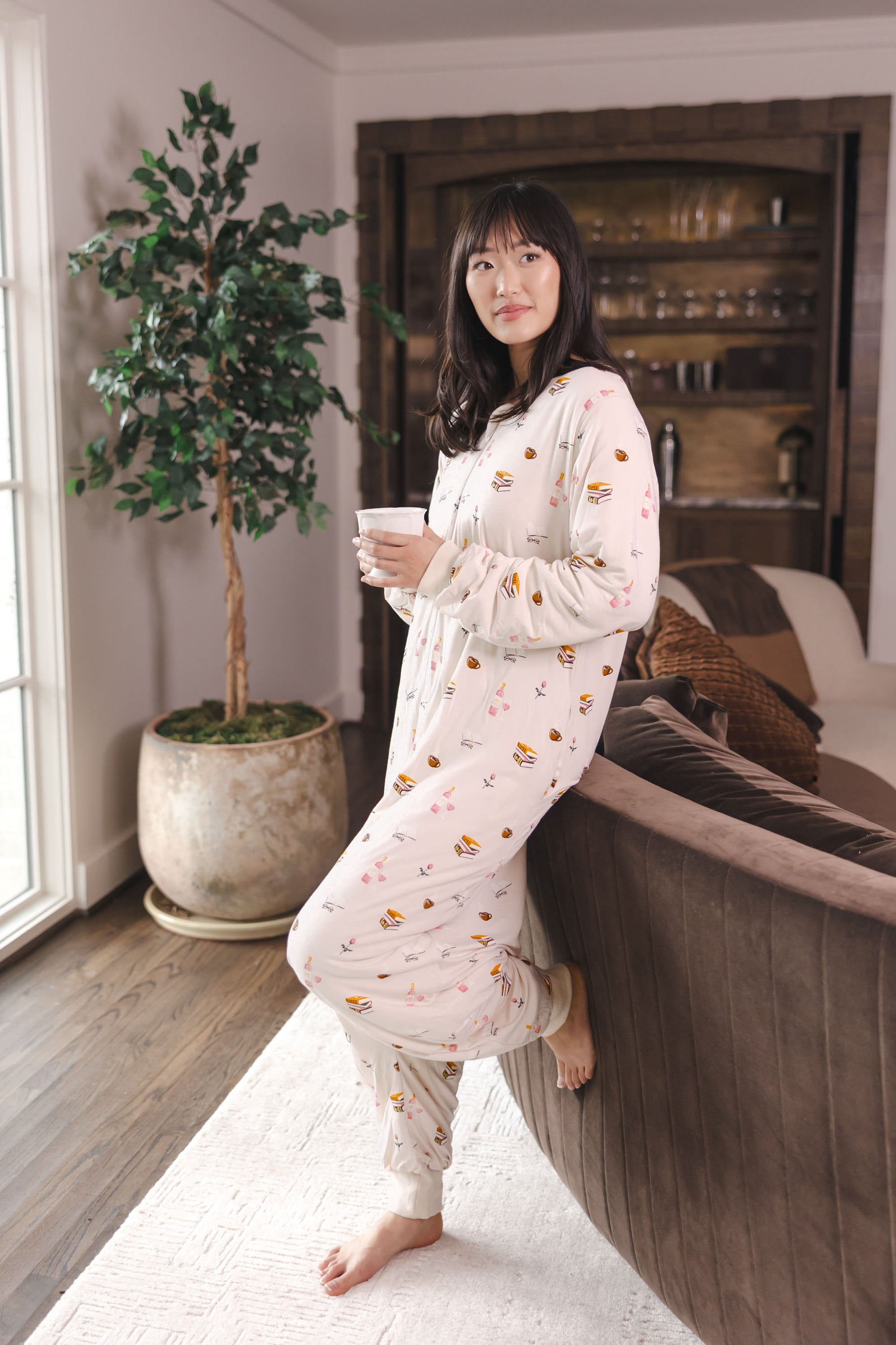 Modal holding a cup in a living room against a sofa wearing Beige Long Sleeve Adult Sleep Bag Walker in Book Club print