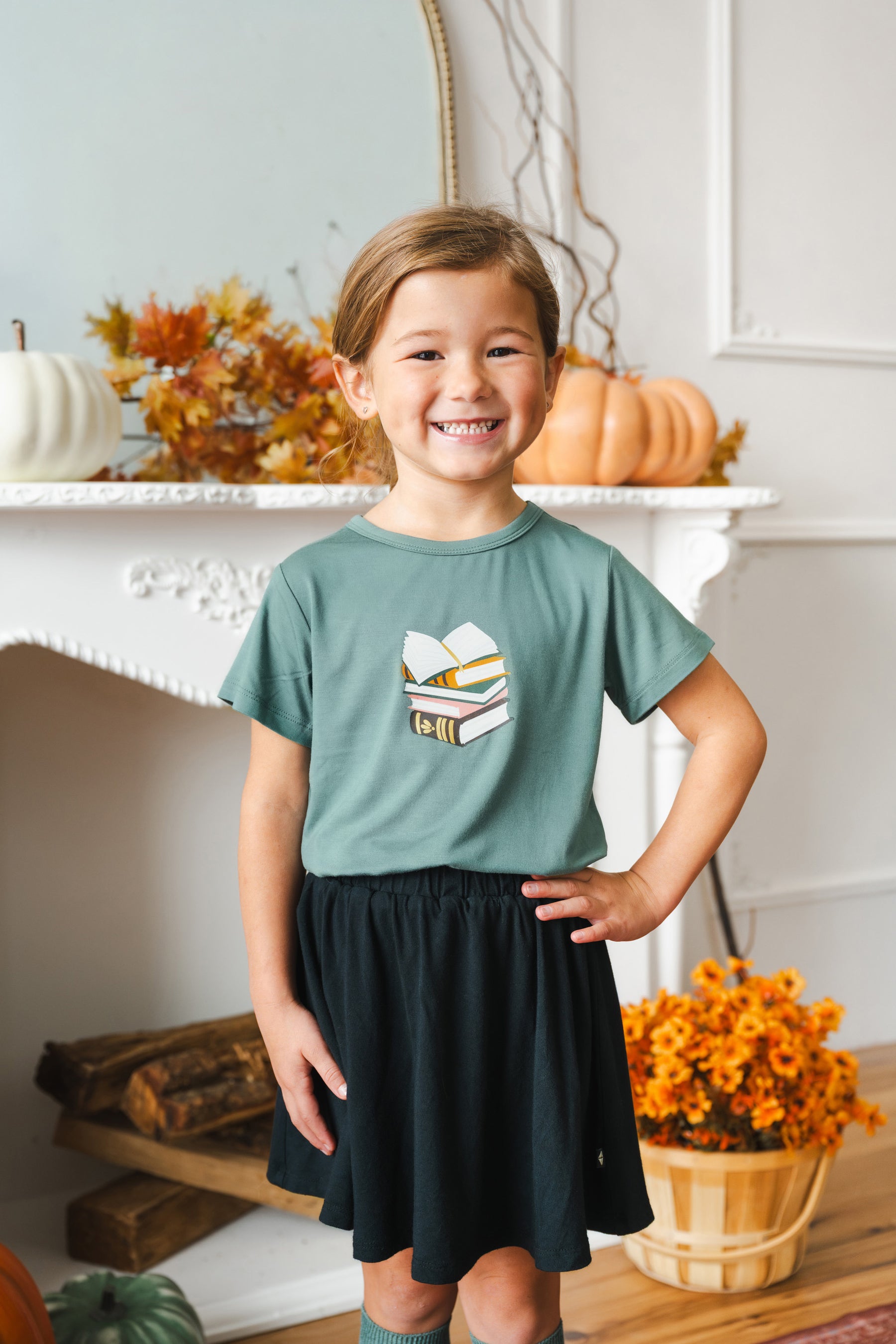 A young girl smiling in a green graphic t-shirt and a black skirt, standing in front of a white fireplace decorated with pumpkins and autumn flowers.