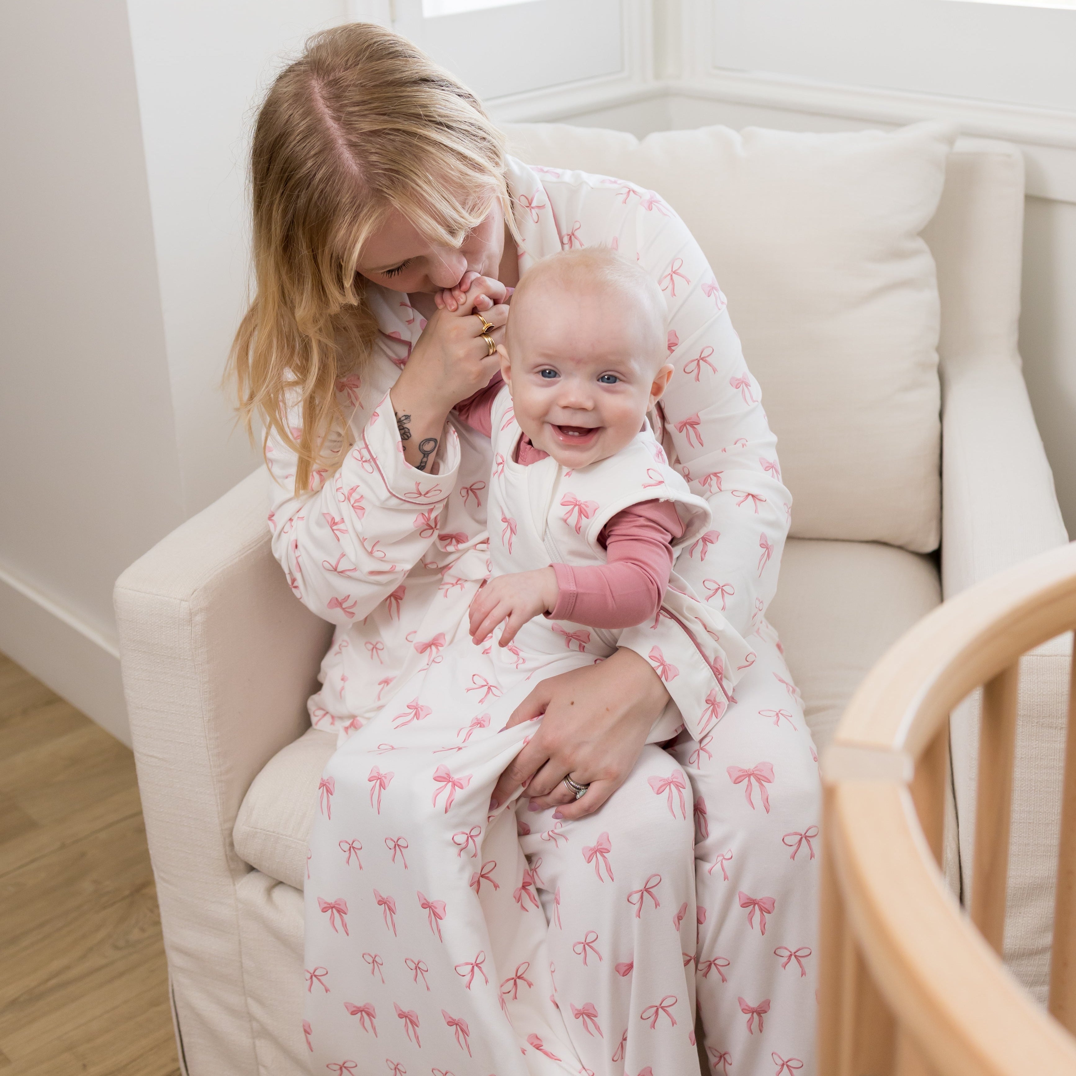 A woman and a baby wearing matching pink bow-patterned sleep sacks sit together in a cream-colored armchair in a bright room.