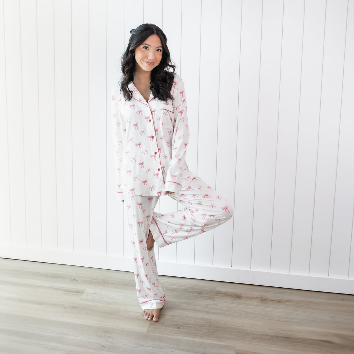 Female standing on one leg with the other leg bent and foot resting on her knee wearing the Long-Sleeved Women's Pajama Set in Bow in front of a white paneled wall