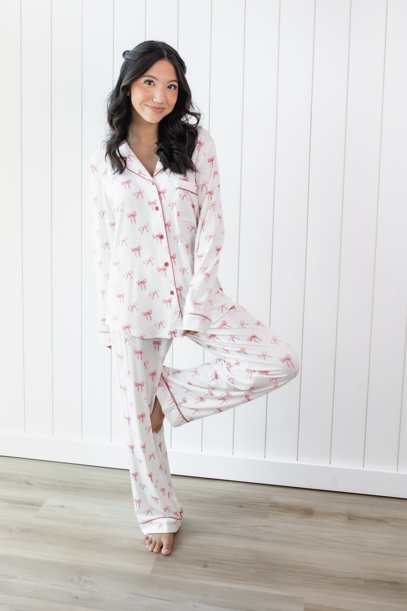 Female standing on one leg with the other leg bent and foot resting on her knee wearing the Long-Sleeved Women's Pajama Set in Bow in front of a white paneled wall
