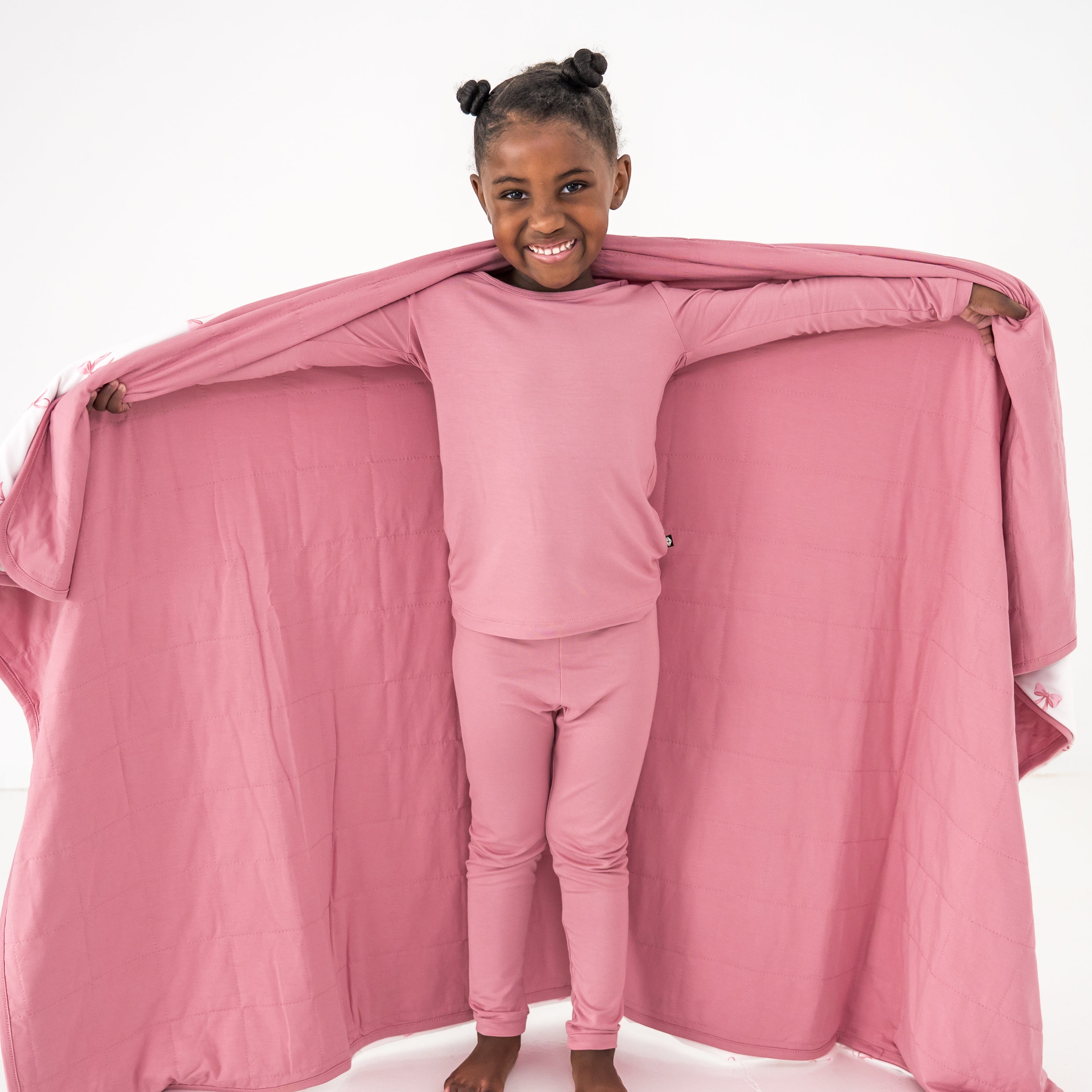 Child in pink pajama set with a pink blanket draped over them on a white background