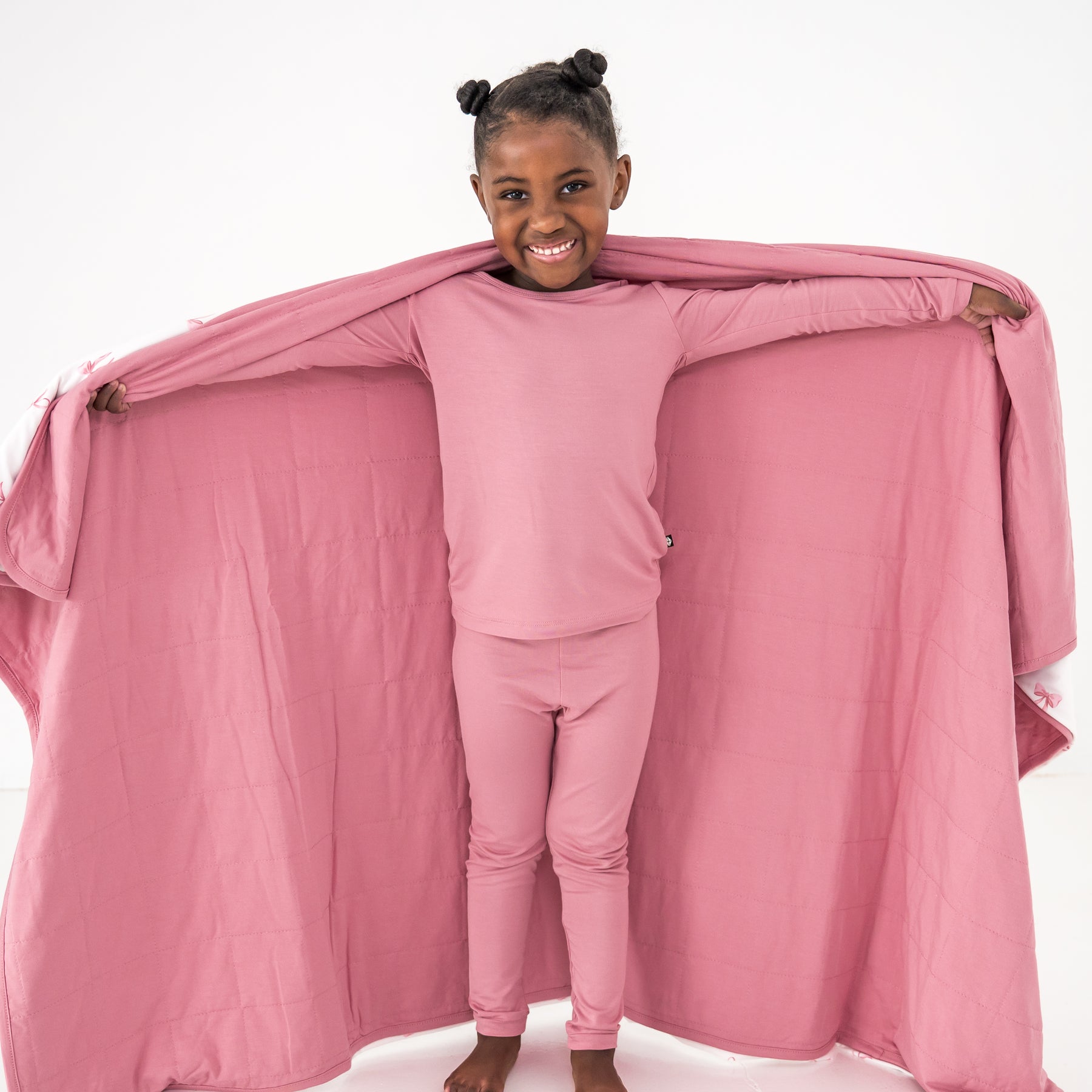 Child in pink pajama set with a pink blanket draped over them on a white background
