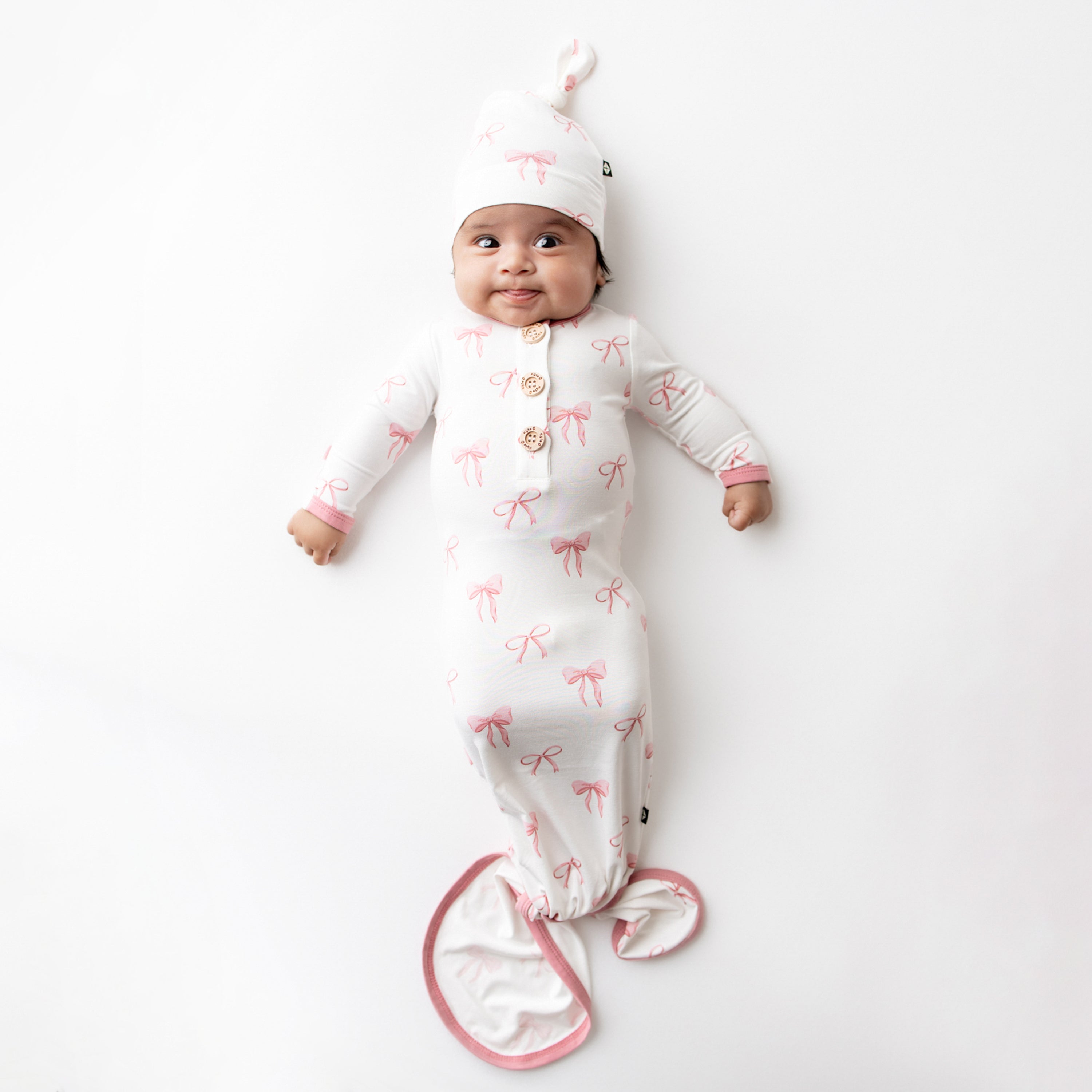 Smiling infant laying on a white surface wearing the Knotted Gown with Hat Set in Bow