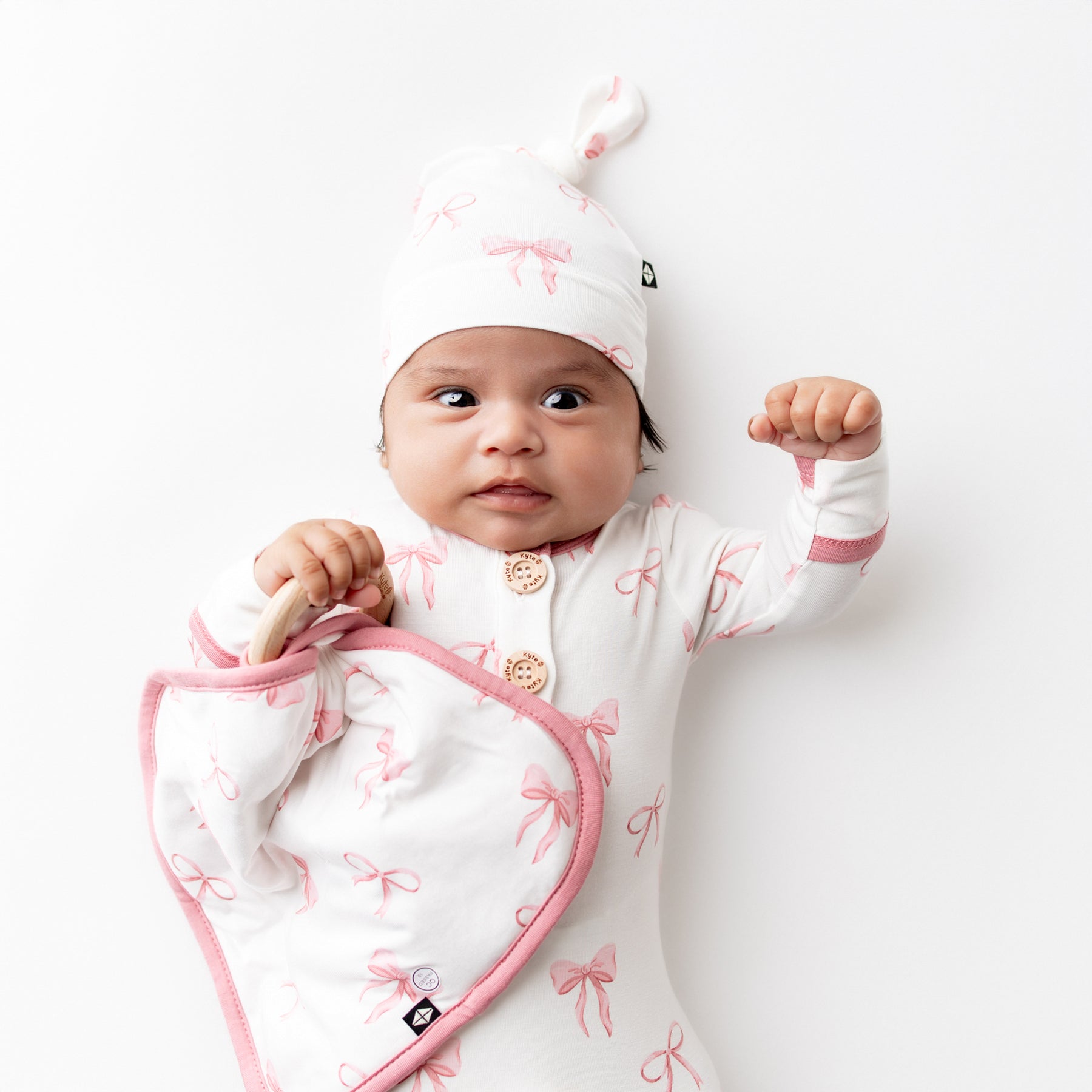 Infant laying on a white surface holding the Lovey in Bow with Removable Wooden Teething Ring wearing a matching knotted gown with hat set