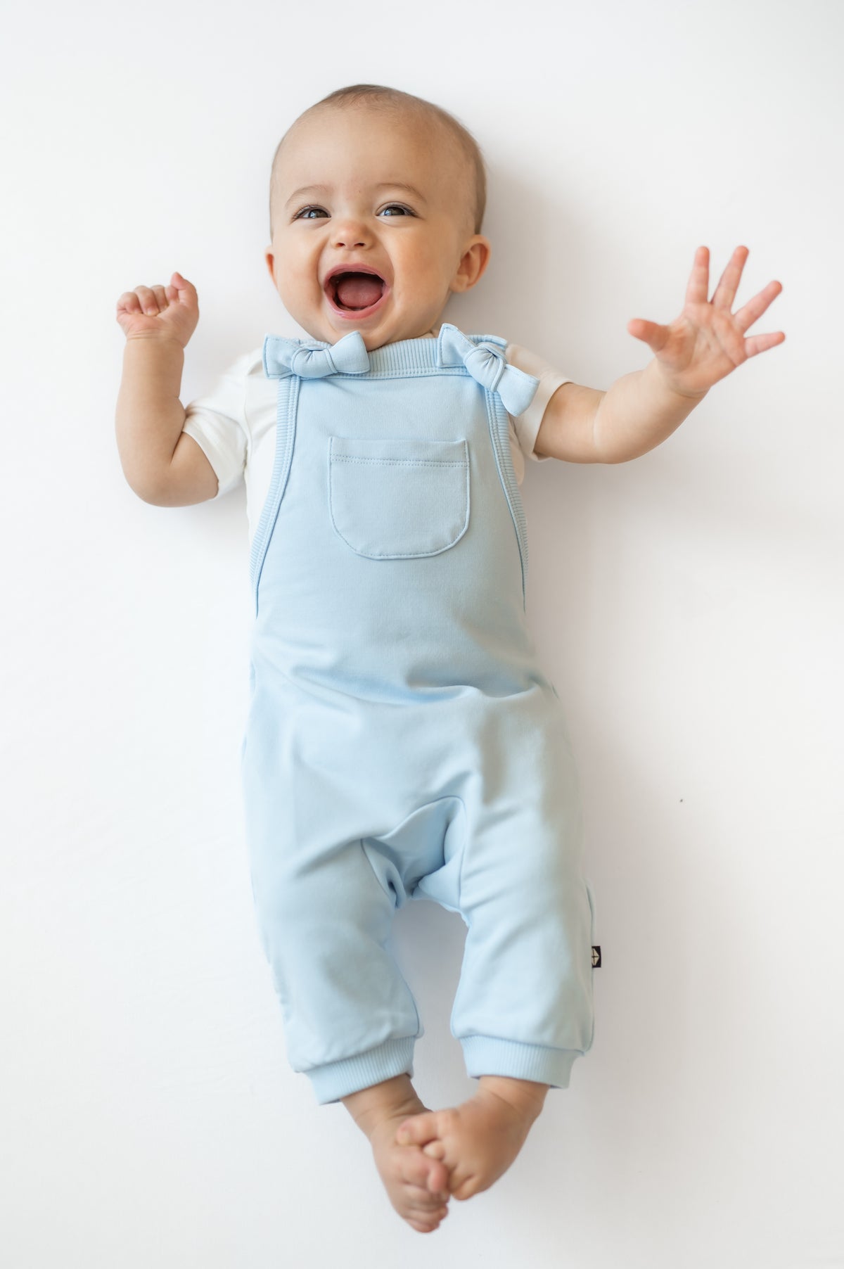 Smiling infant wearing the Overall in Breeze on a light colored surface