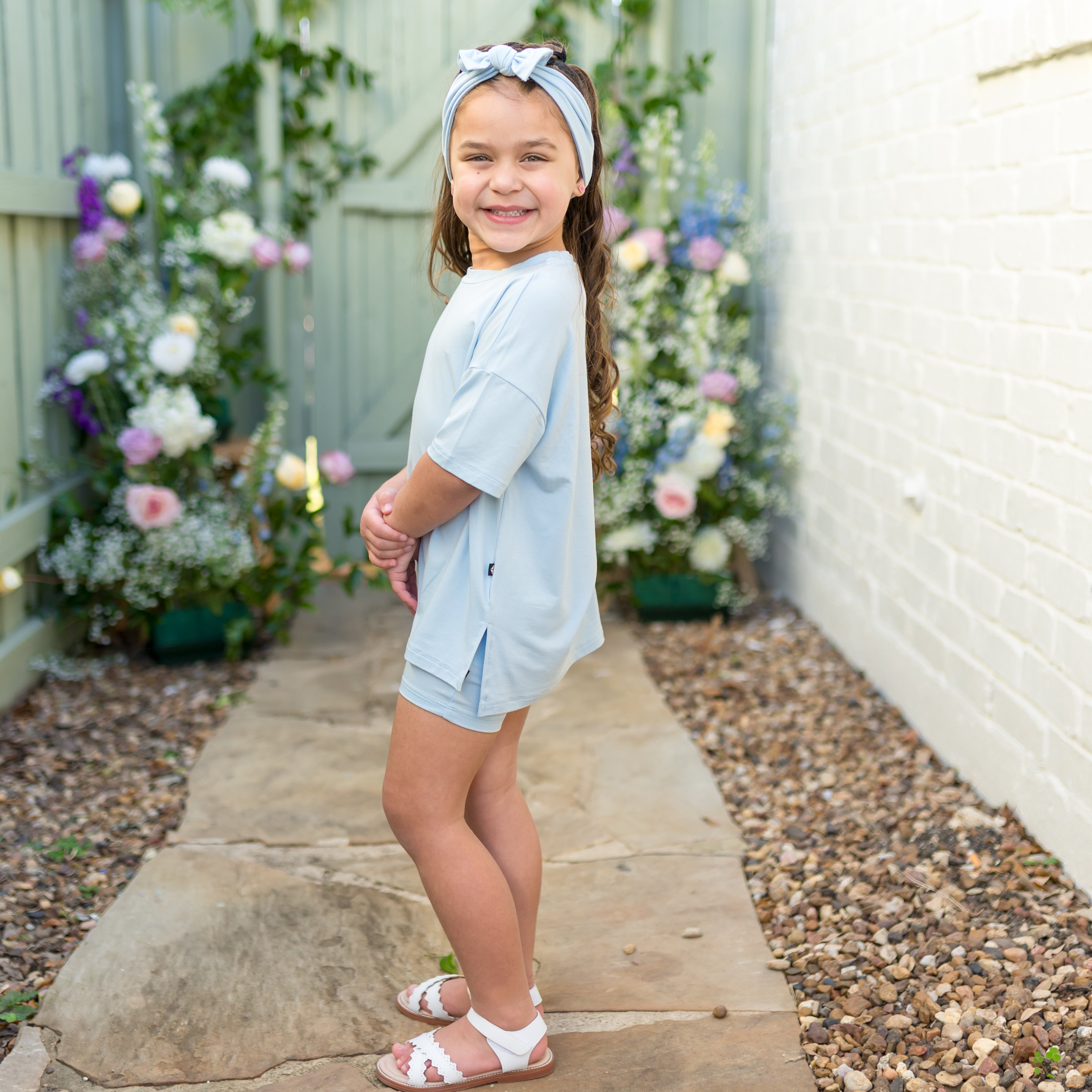 Young girl in light blue outfit standing outdoors with flowers and a white wall in the background