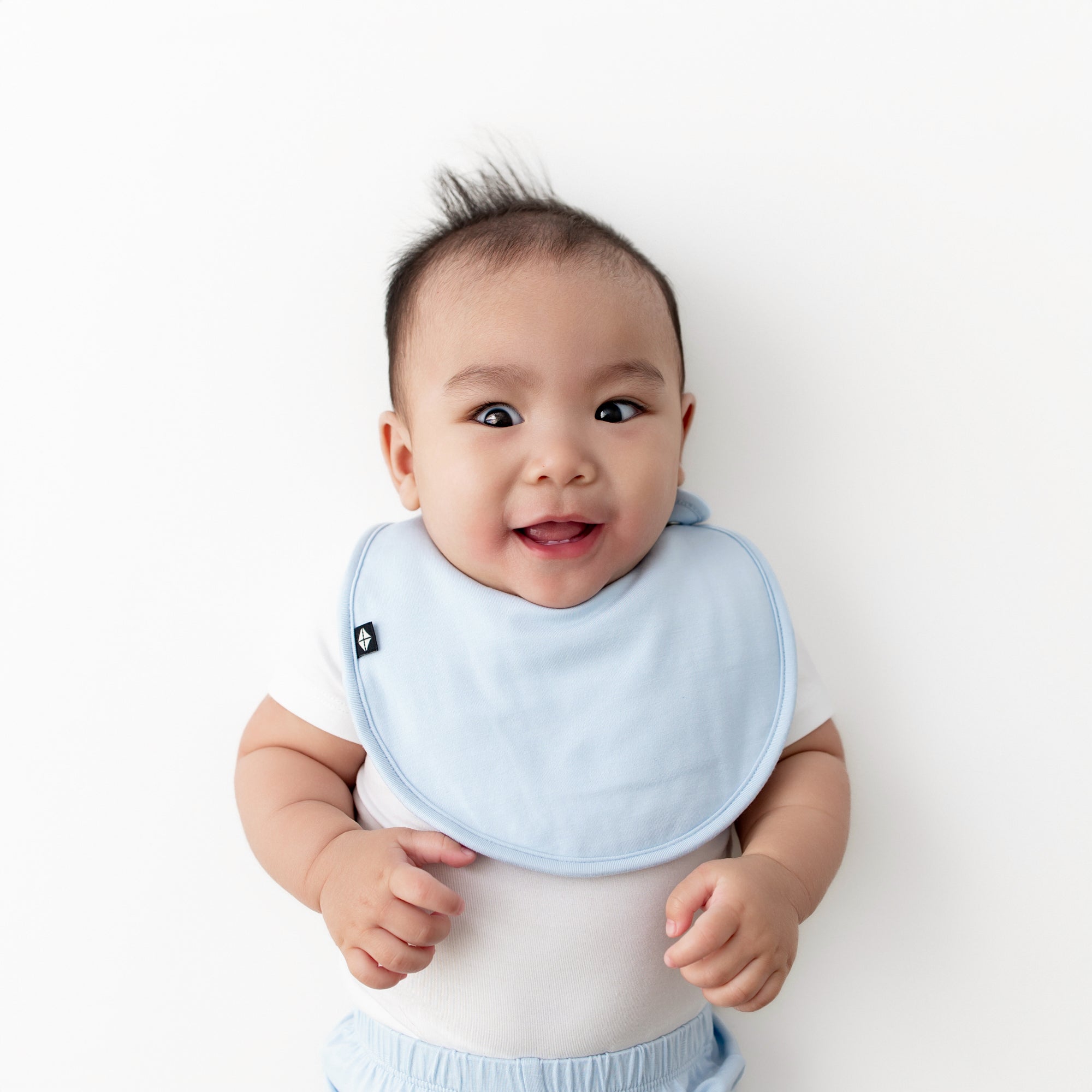 Infant laying on a light neutral surface wearing the Cushy Bib in Breeze