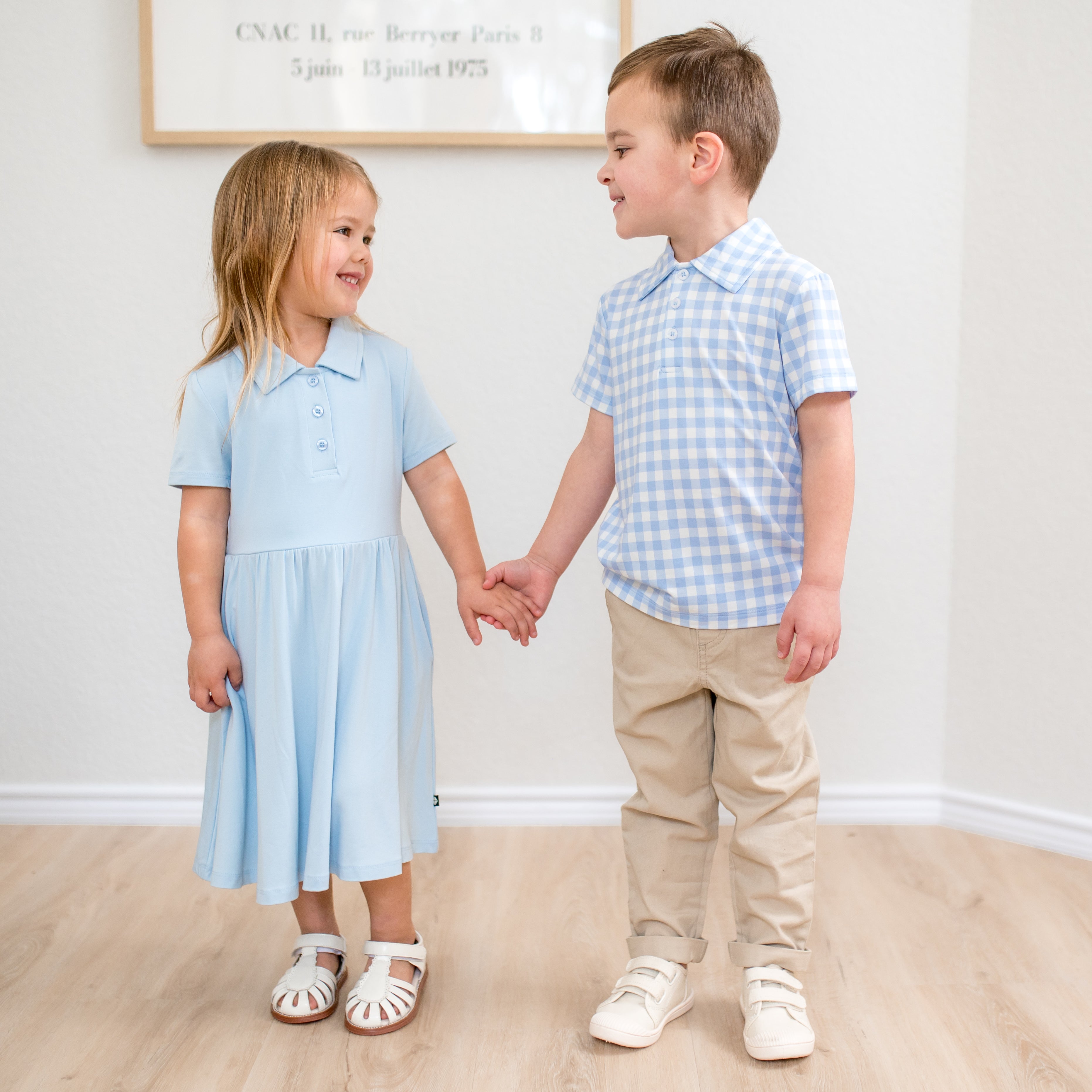 Brother wearing the Toddler Short Sleeve Polo in Gingham Breeze with khaki colored pants holding his sisters hand who is wearing a Breeze polo dress