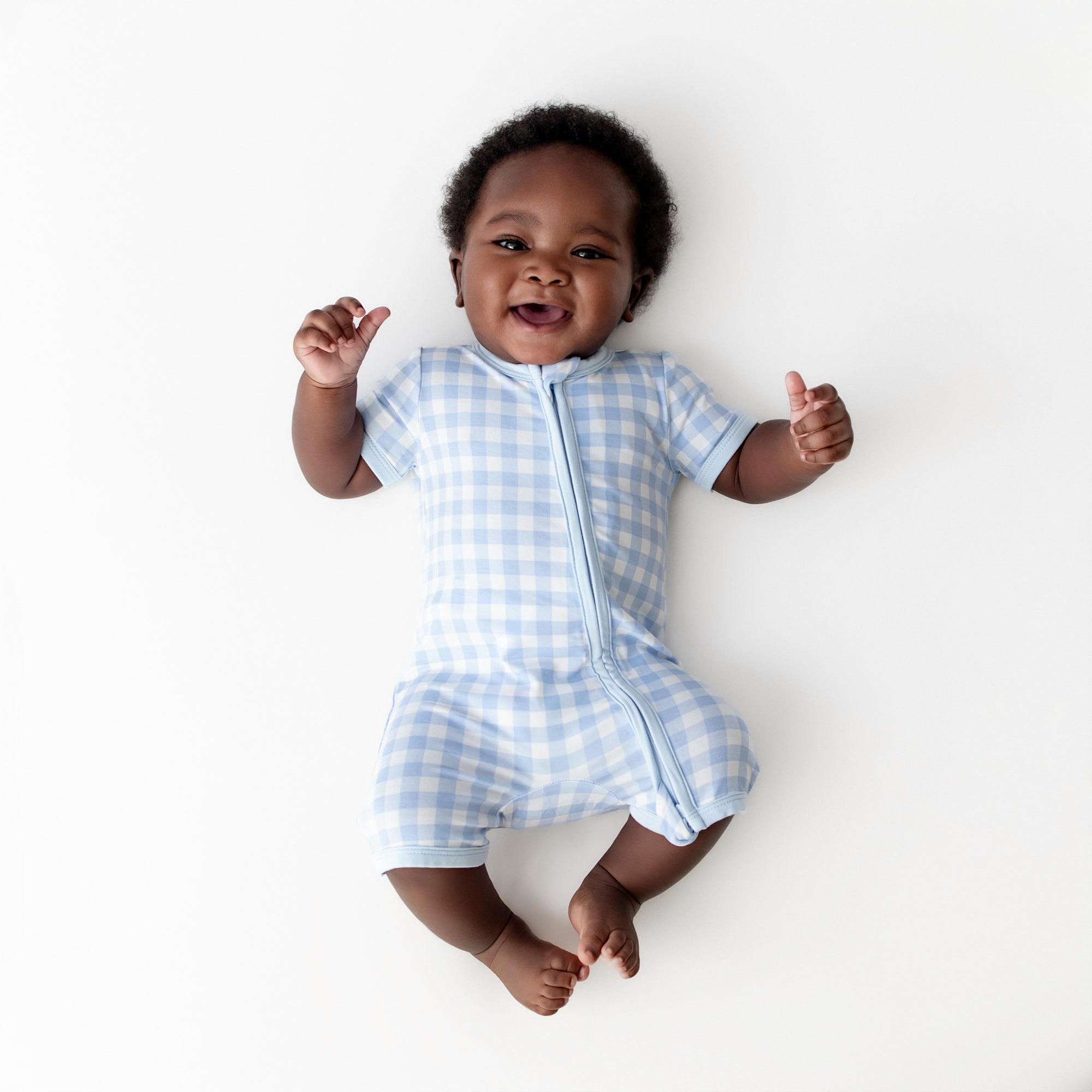 Young infant wearing the Zipper Shortall Romper in Gingham Breeze laying on a light cream surface