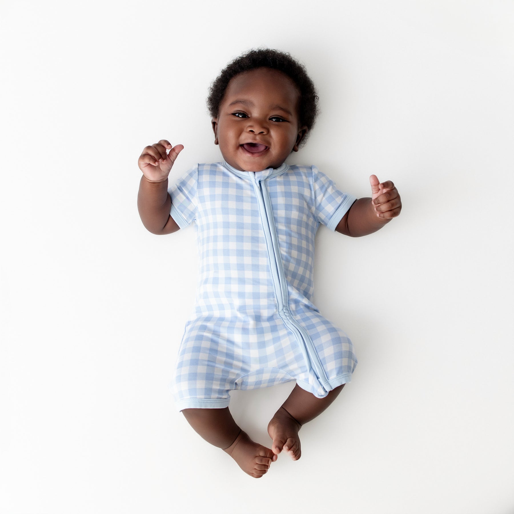 Young infant wearing the Zipper Shortall Romper in Gingham Breeze laying on a light cream surface