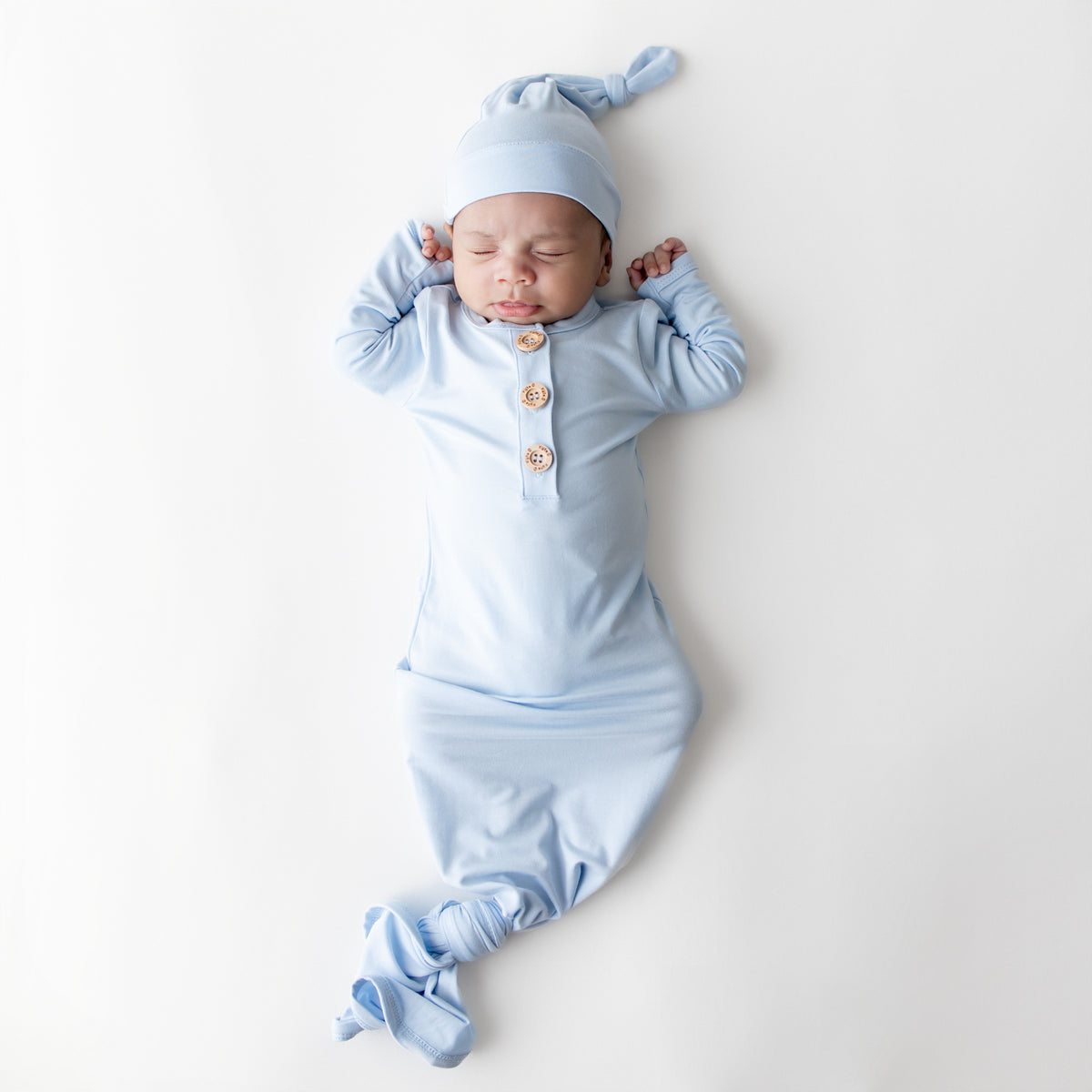 Sleeping newborn wearing the Knotted Gown with Hat Set in Breeze laying on  a white surface