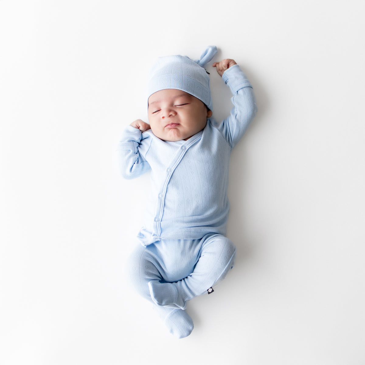 Sleeping newborn laying on a white surface wearing the Pointelle Take Me Home Set with Cap in Breeze