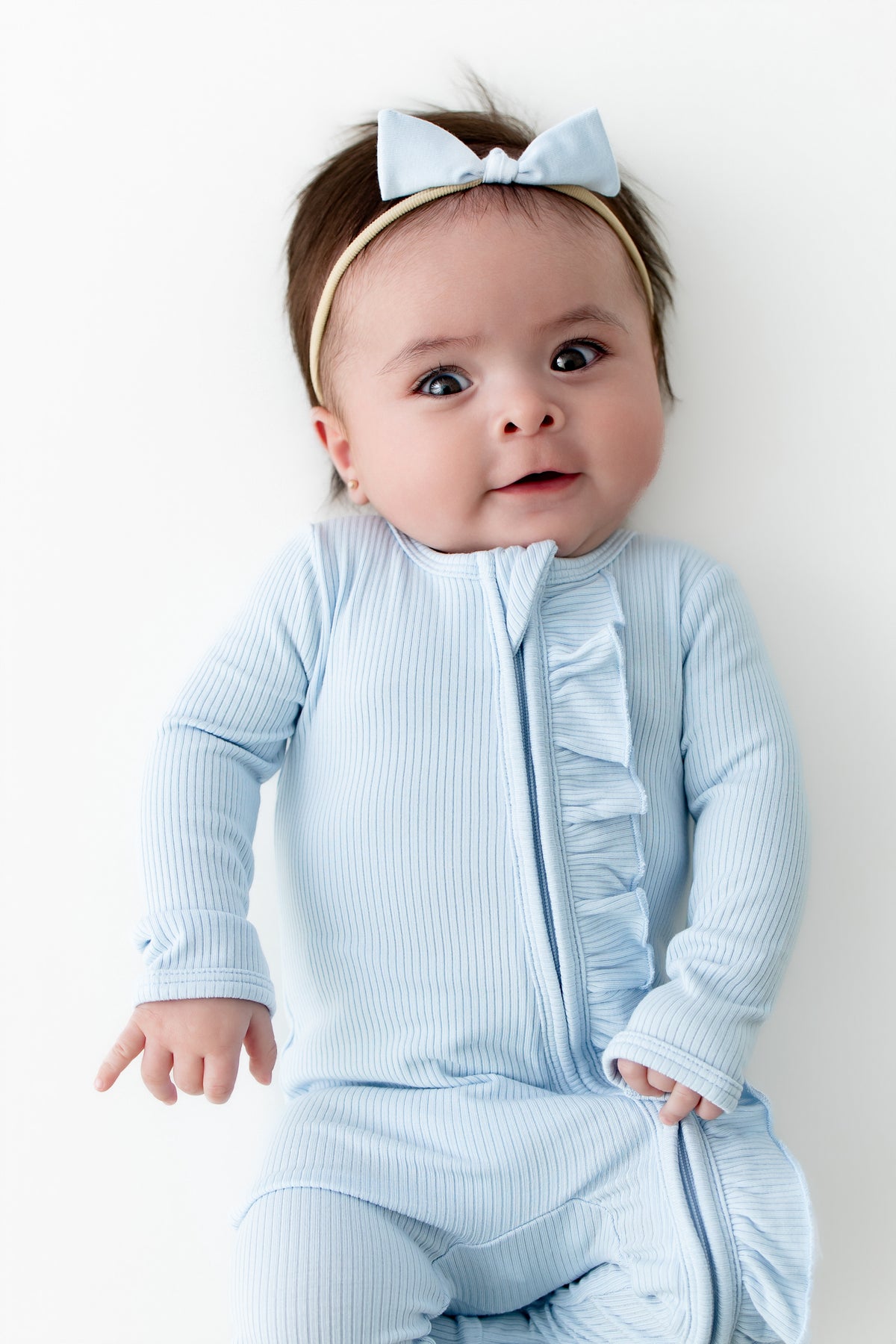 Infant girl wearing the Ribbed Ruffle Zipper Footie in Breeze with matching tiny nylon bow