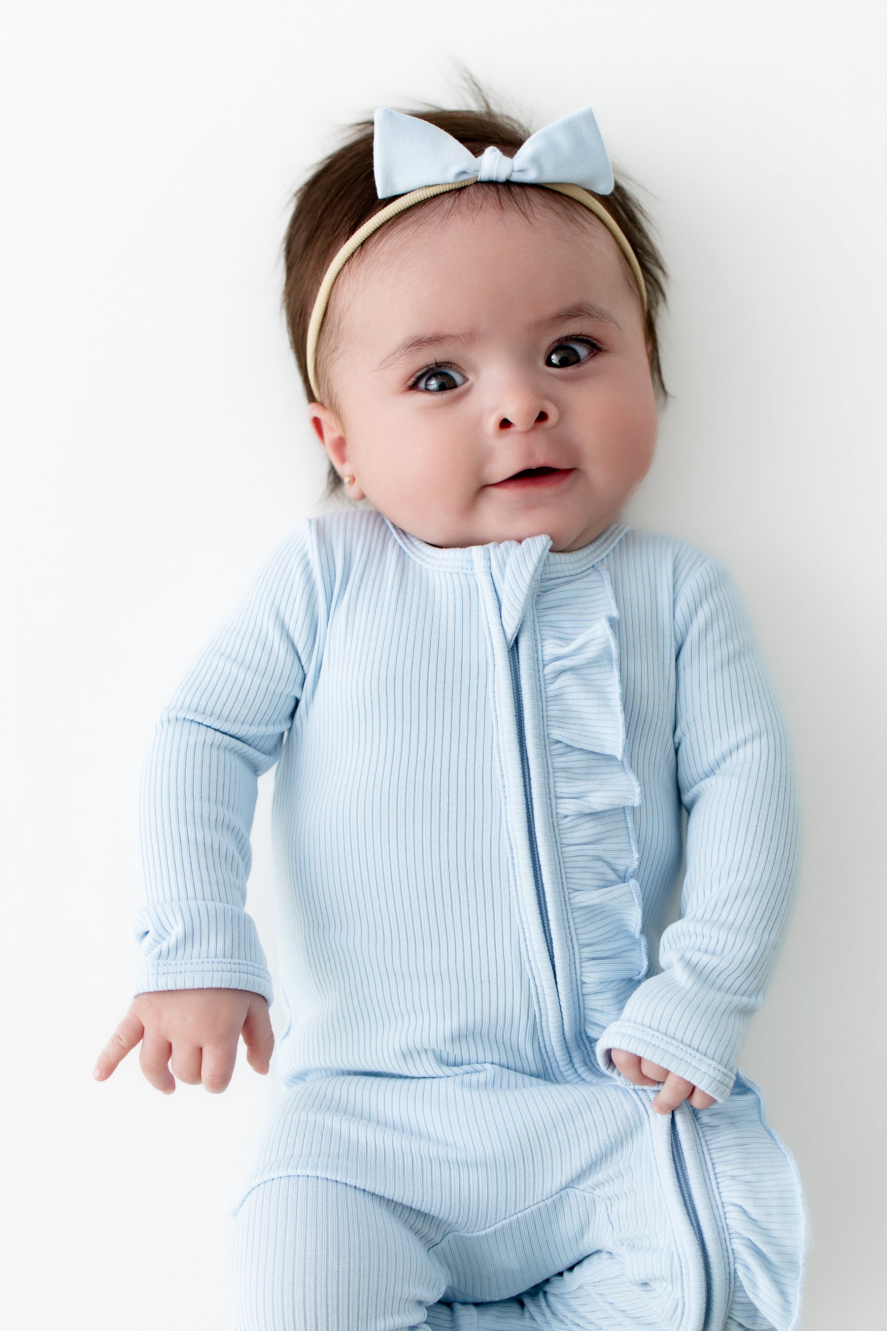 Infant girl wearing the Ribbed Ruffle Zipper Footie in Breeze with matching tiny nylon bow