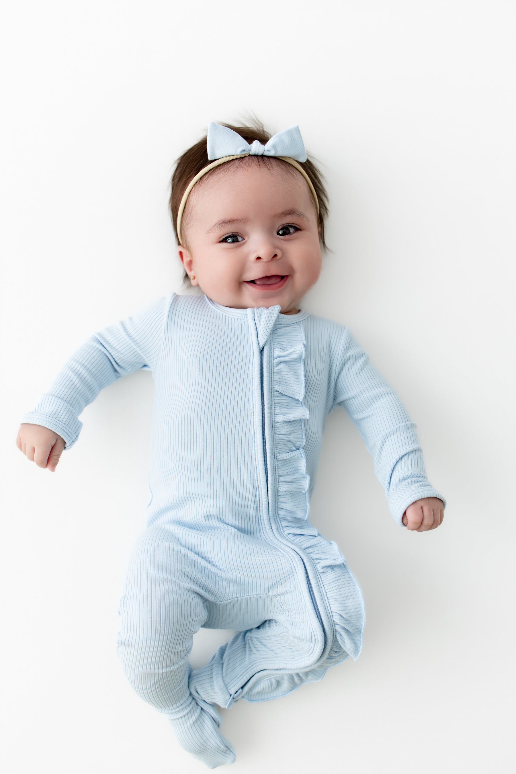 Infant girl laying on a white surface wearing the Ribbed Ruffle Zipper Footie in Breeze with matching tiny nylon bow