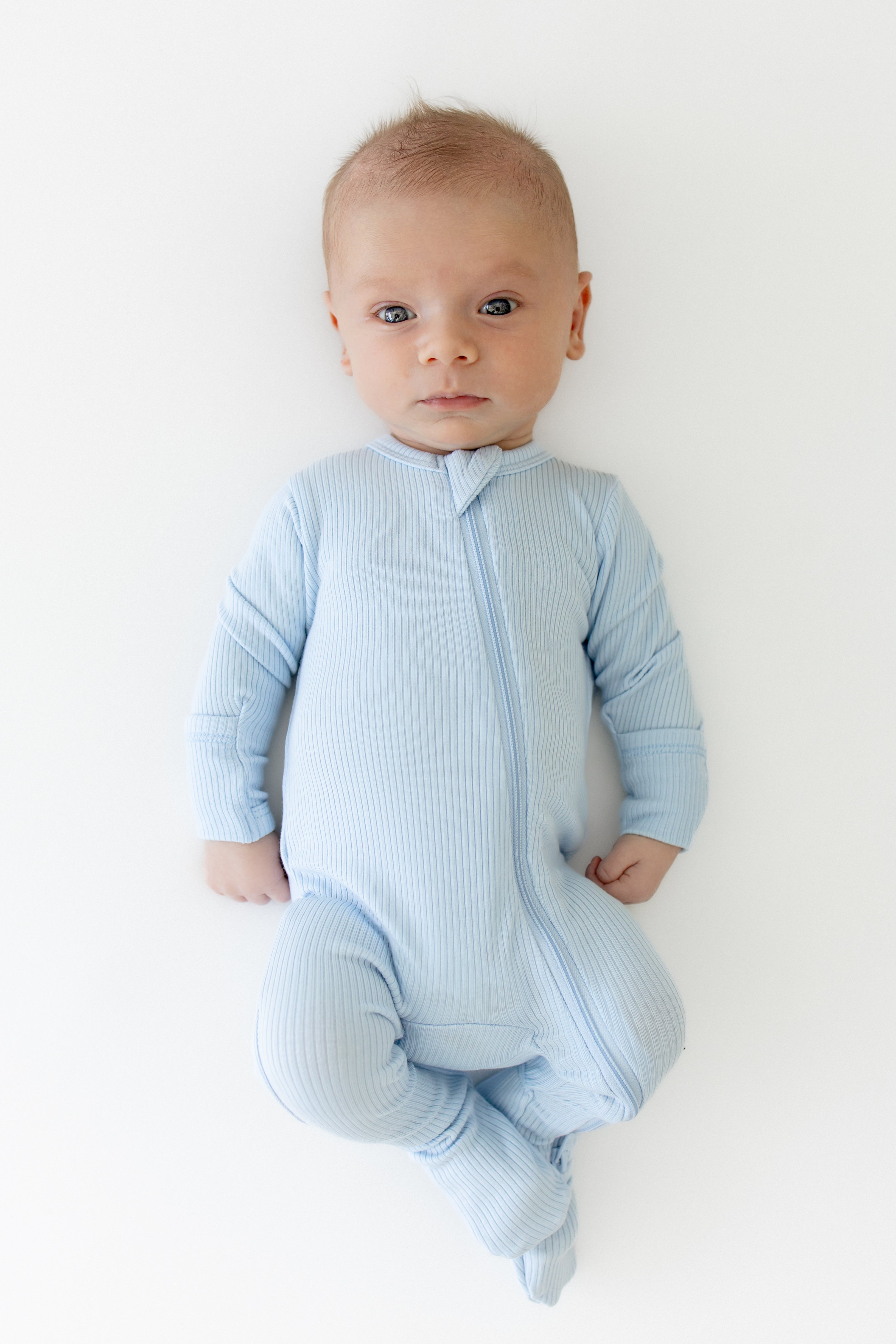 Infant laying on a white surface wearing the Ribbed Zipper Footie in Breeze