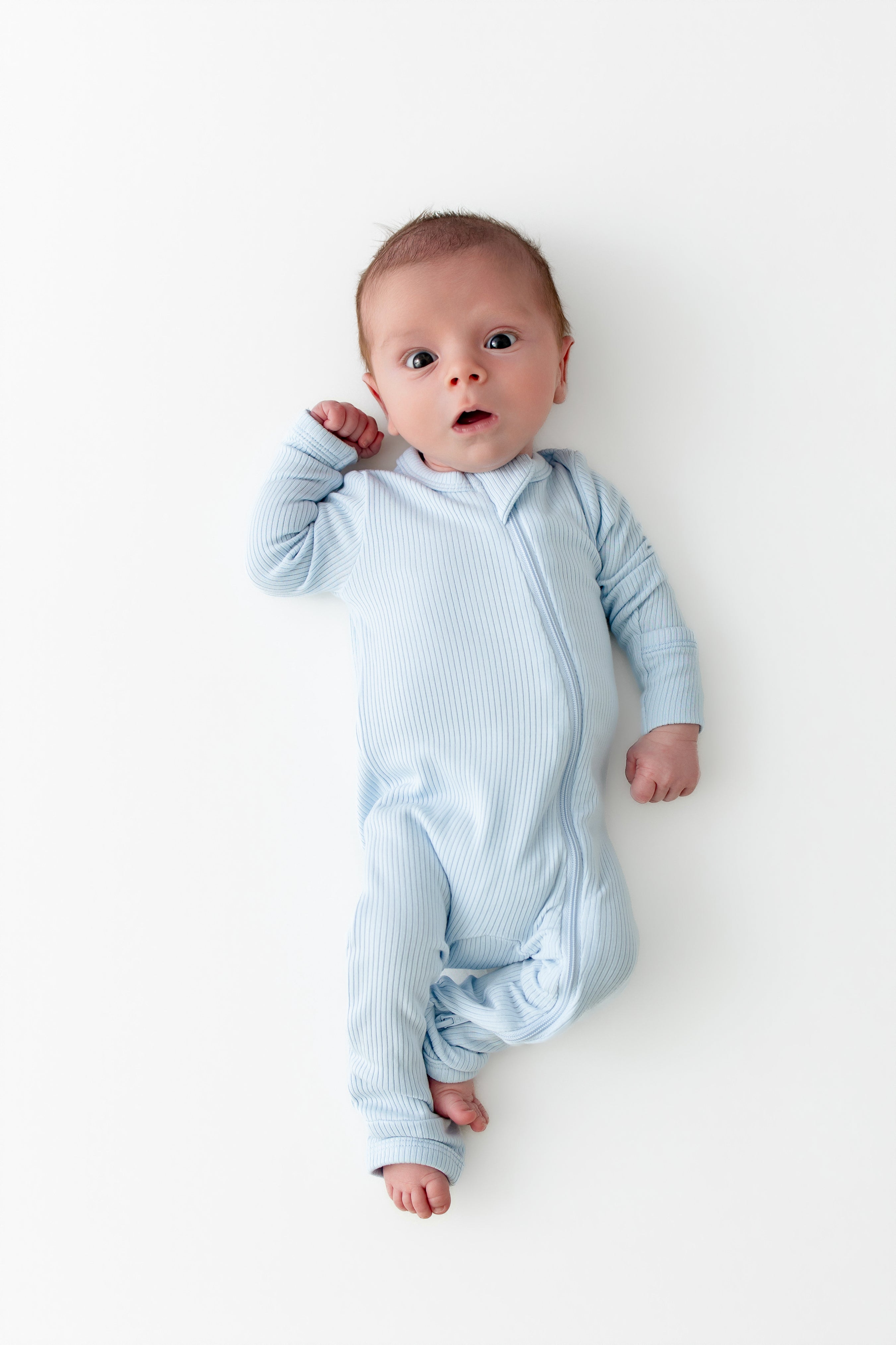 Newborn laying on a white surface wearing the Ribbed Zipper Romper in Breeze
