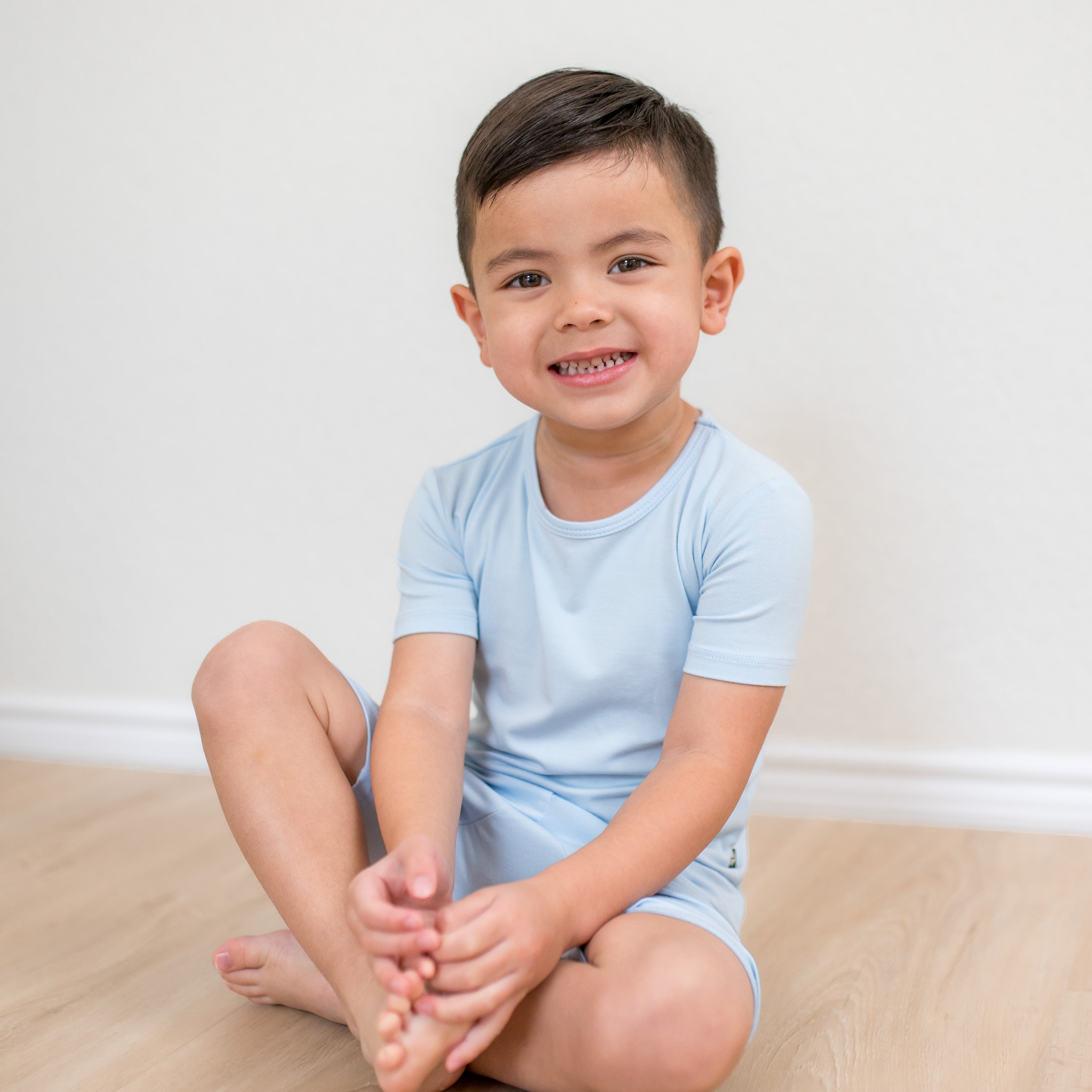 Young boy sitting on a wood grained floor wearing the Short Sleeve Pajamas in Breeze