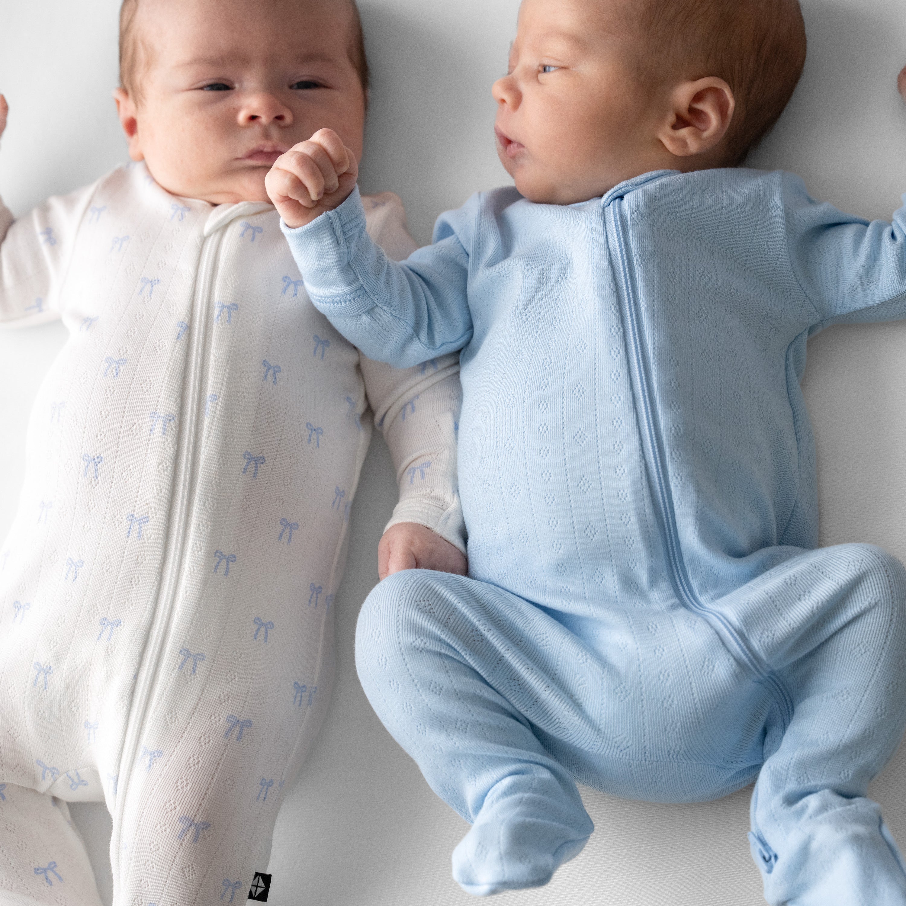 Two newborn laying side by side wearing the Pointelle Zippered footies in Breeze and Blue bows