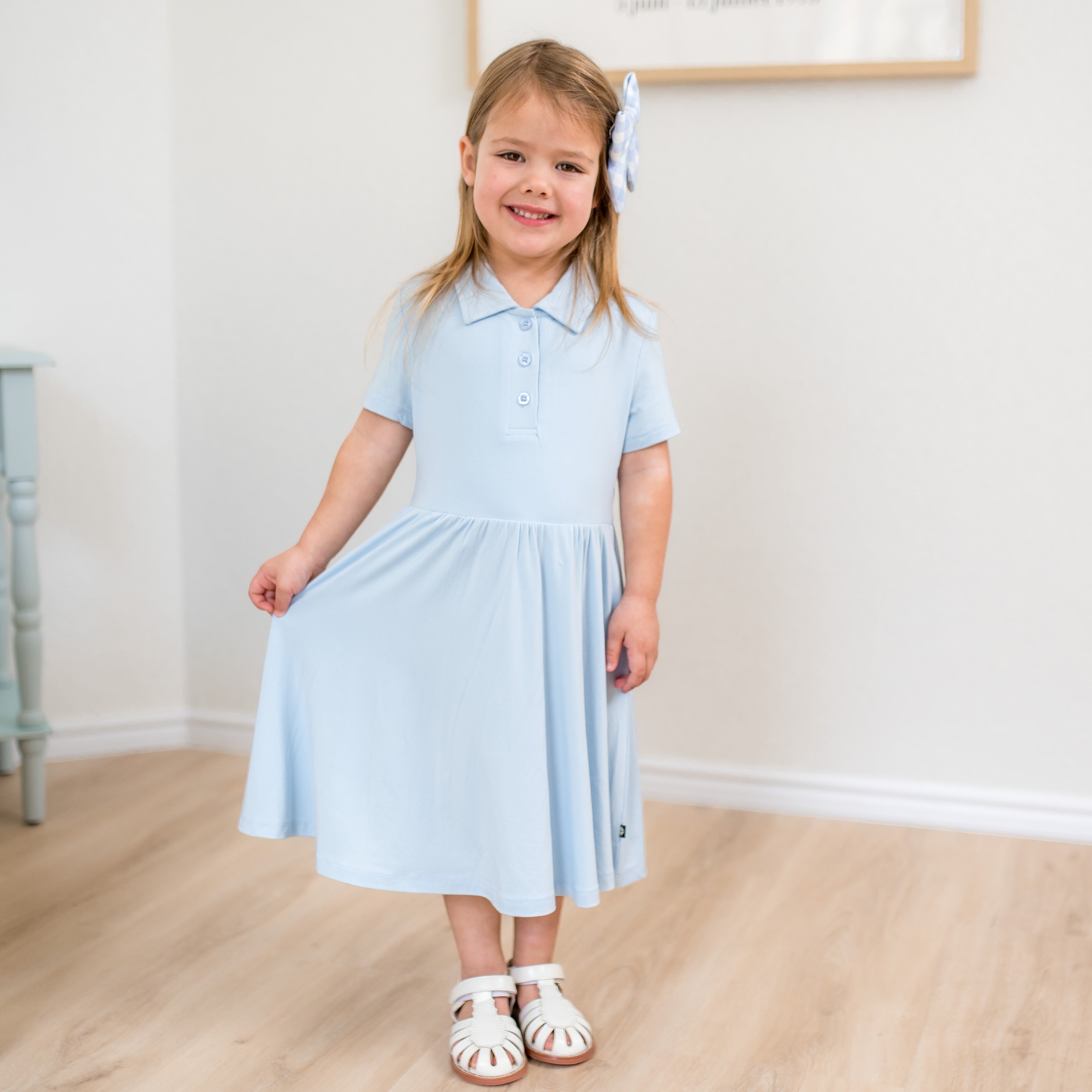 Young girl wearing a light blue polo dress standing indoors with a white wall background