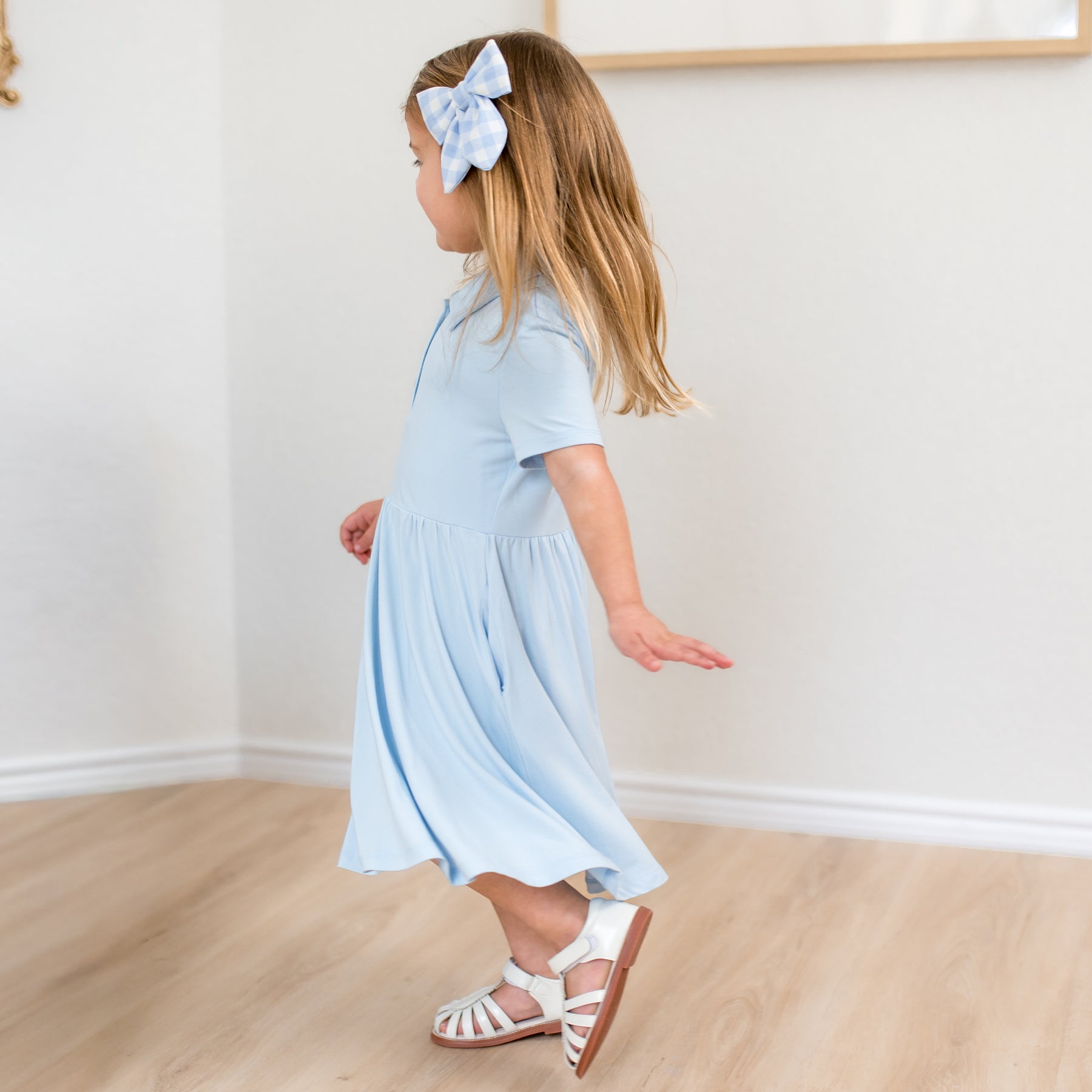 Young girl in a light blue dress with a bow in her hair standing on a wooden floor.