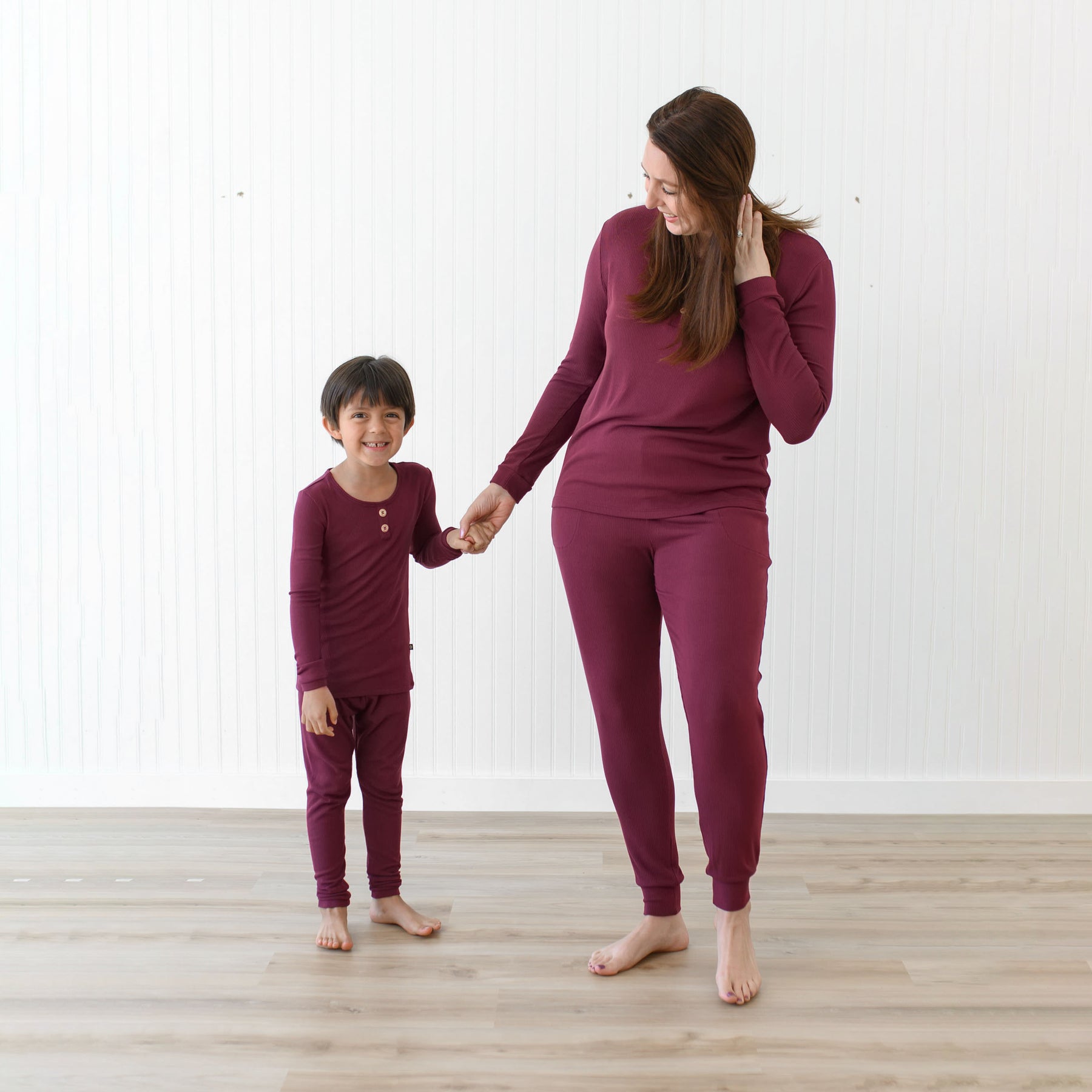 Mother standing beside her son holding his hand looking at him both wearing the Ribbed Henley Set in Burgundy in front of a white paneled wall