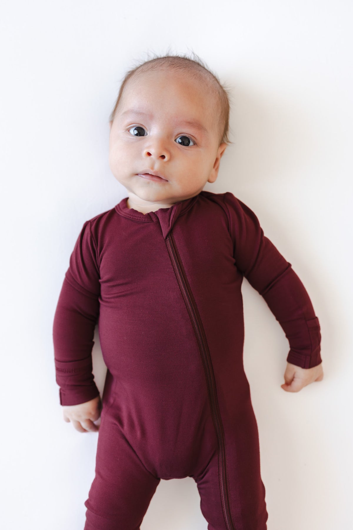 A baby wearing a long-sleeve, one-piece burgundy zip-up romper, lying on a plain white background.