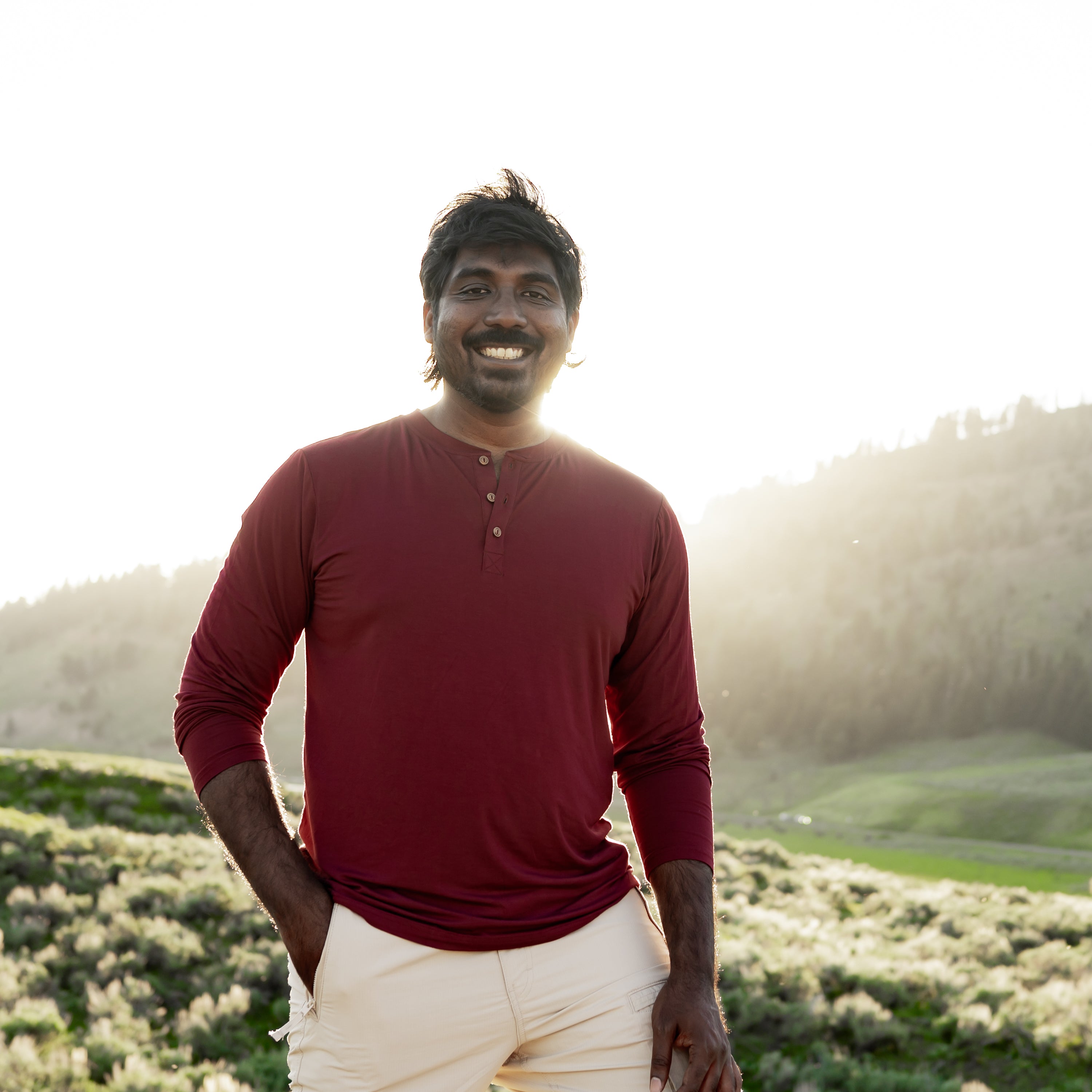 Smiling male model standing in a field wearing the Men's Long Sleeve Henley Top in Burgundy
